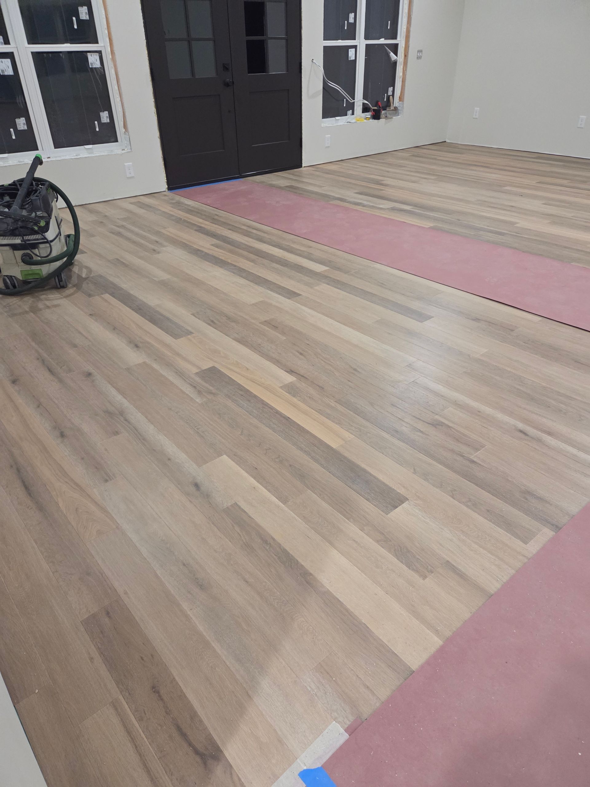 A room under renovation with light-toned hardwood floors partially installed over a pink underlayment and a floor sander.