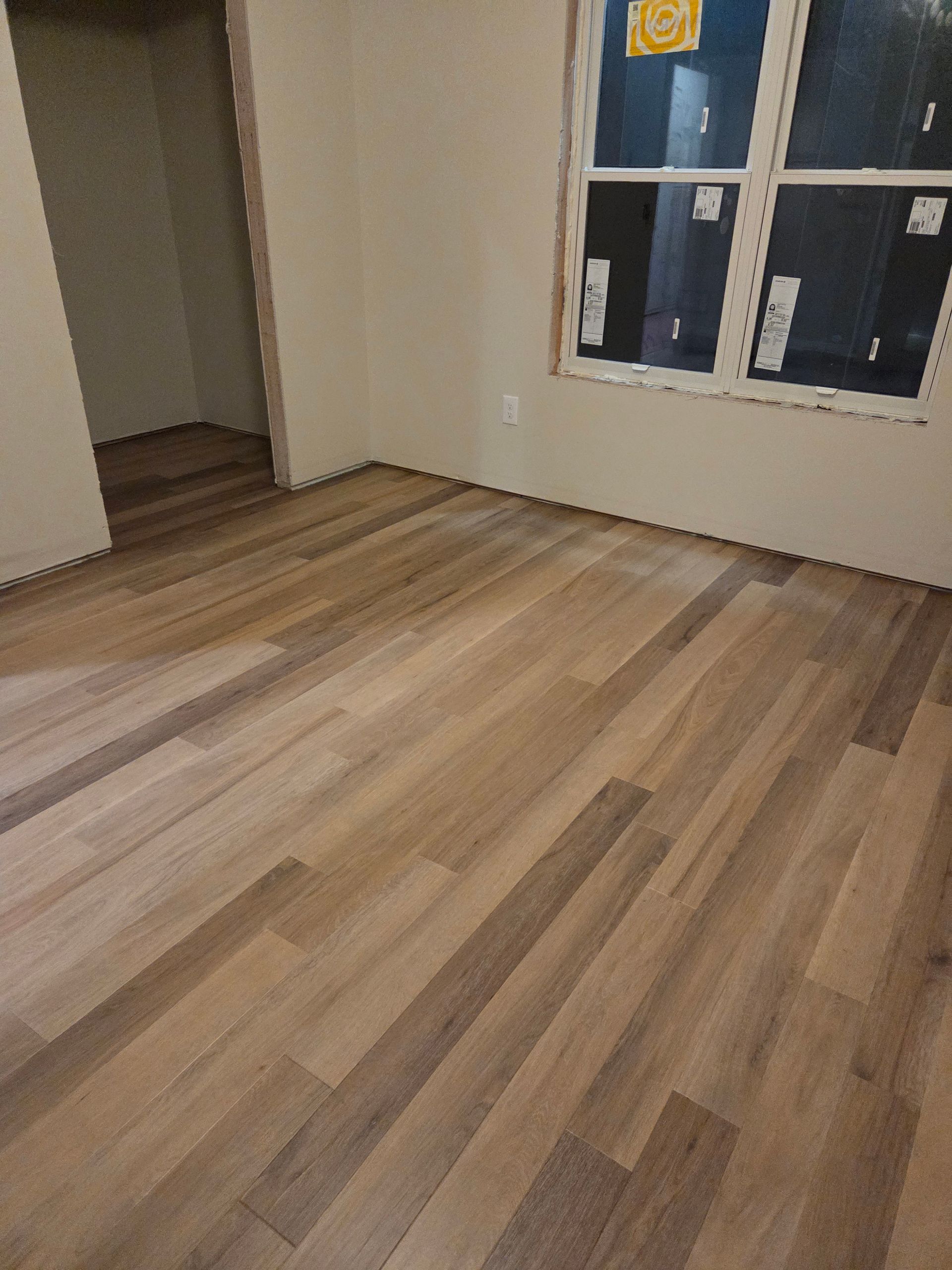 An empty room with light wood plank flooring, neutral walls, and a large window.