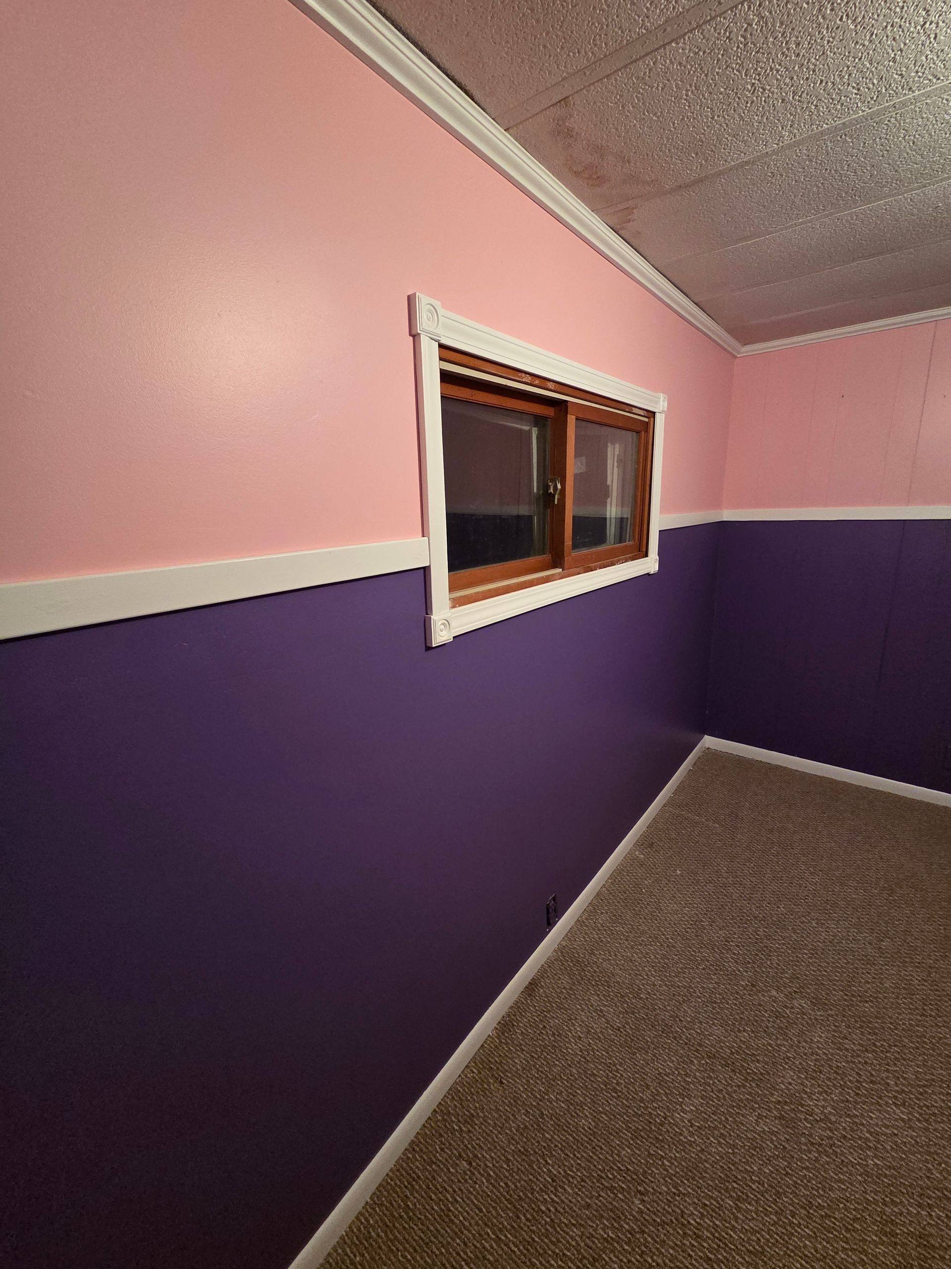 A corner of a room with pink upper walls, purple lower walls separated by white trim, a small window, and textured carpet.