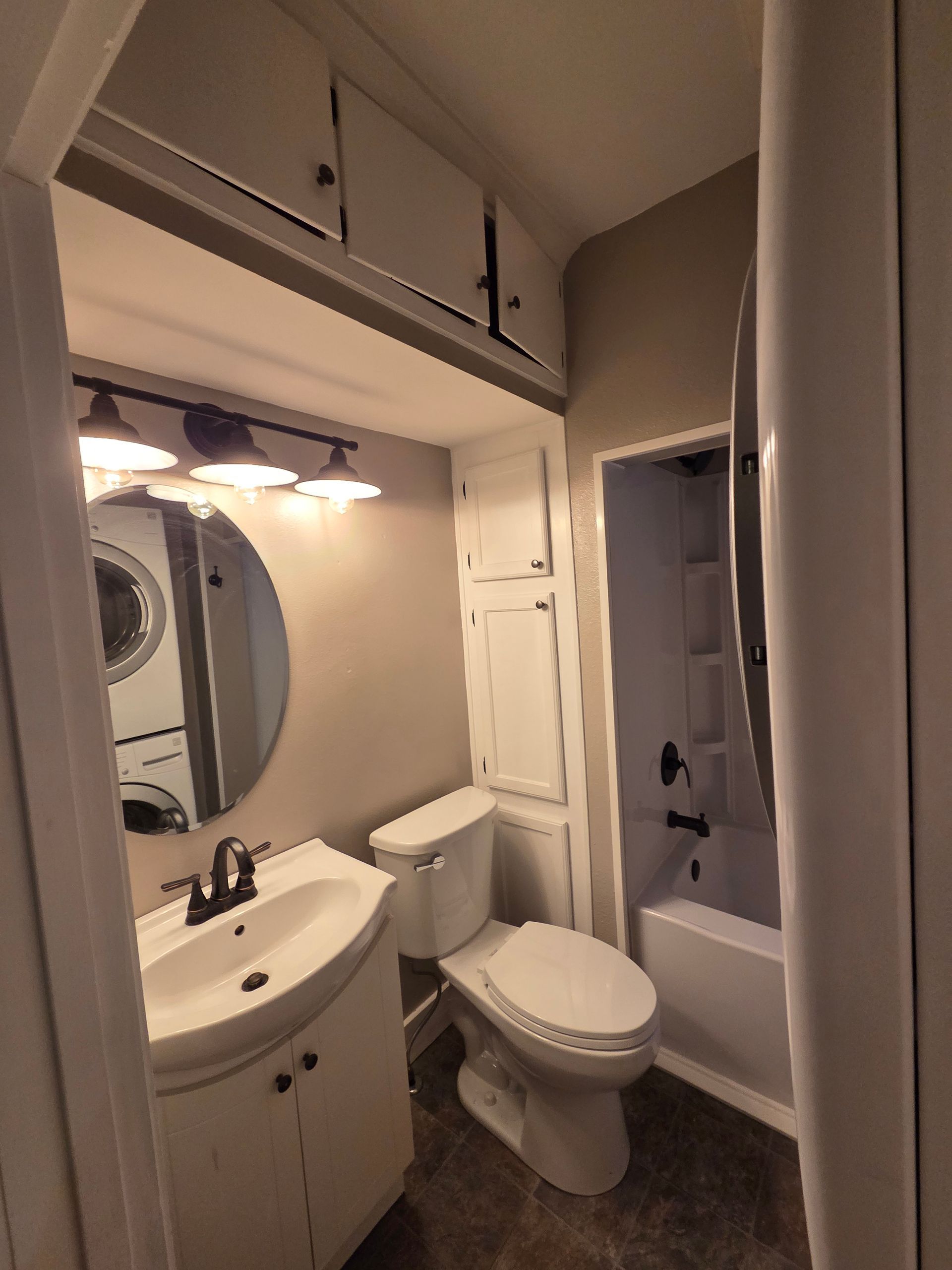 A small bathroom featuring a vanity with a round mirror, a white toilet, and a bathtub with a shower head.