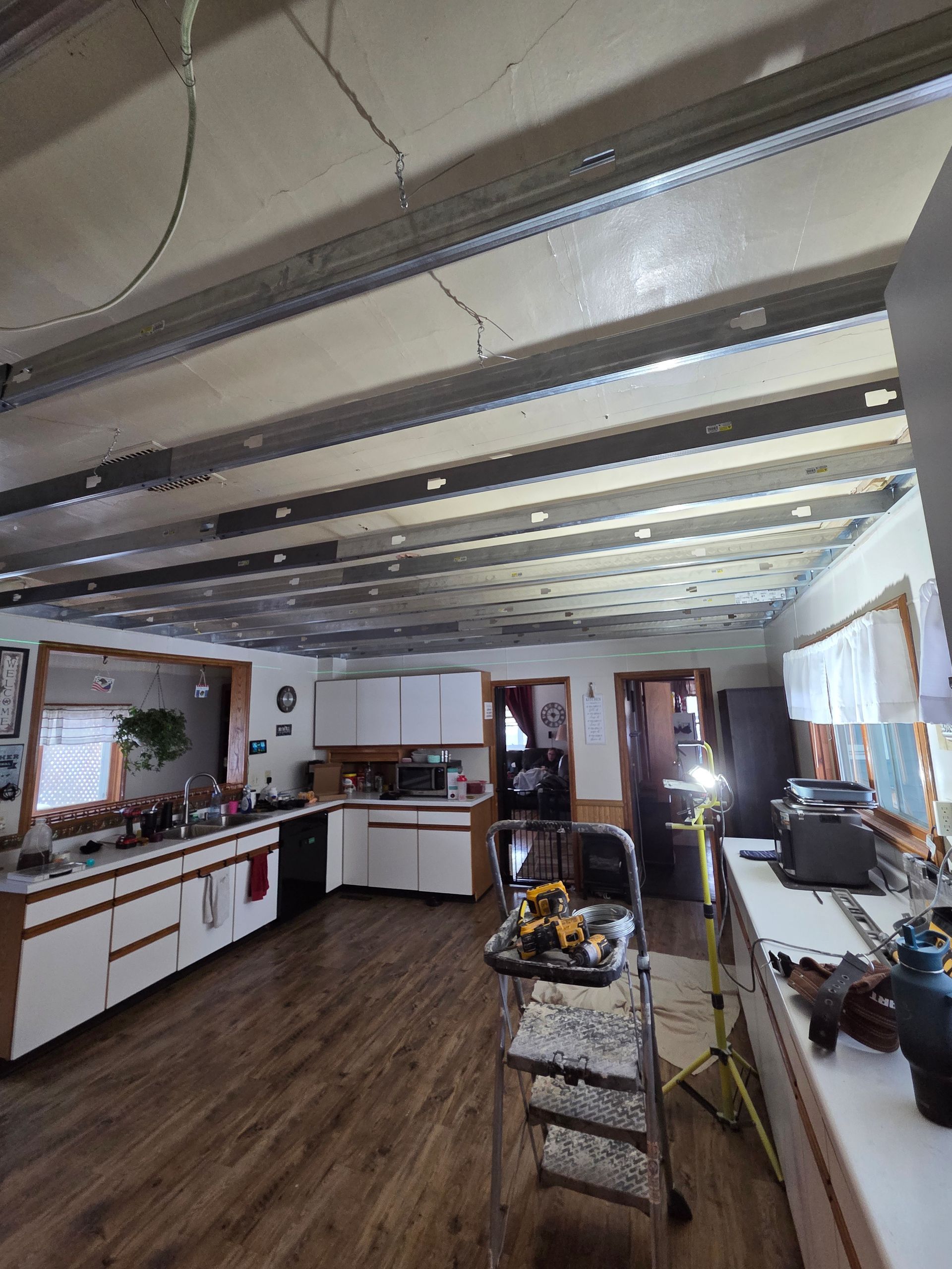 A kitchen interior with wooden floors, white cabinets, and an exposed ceiling structure, featuring a step ladder mid-room.