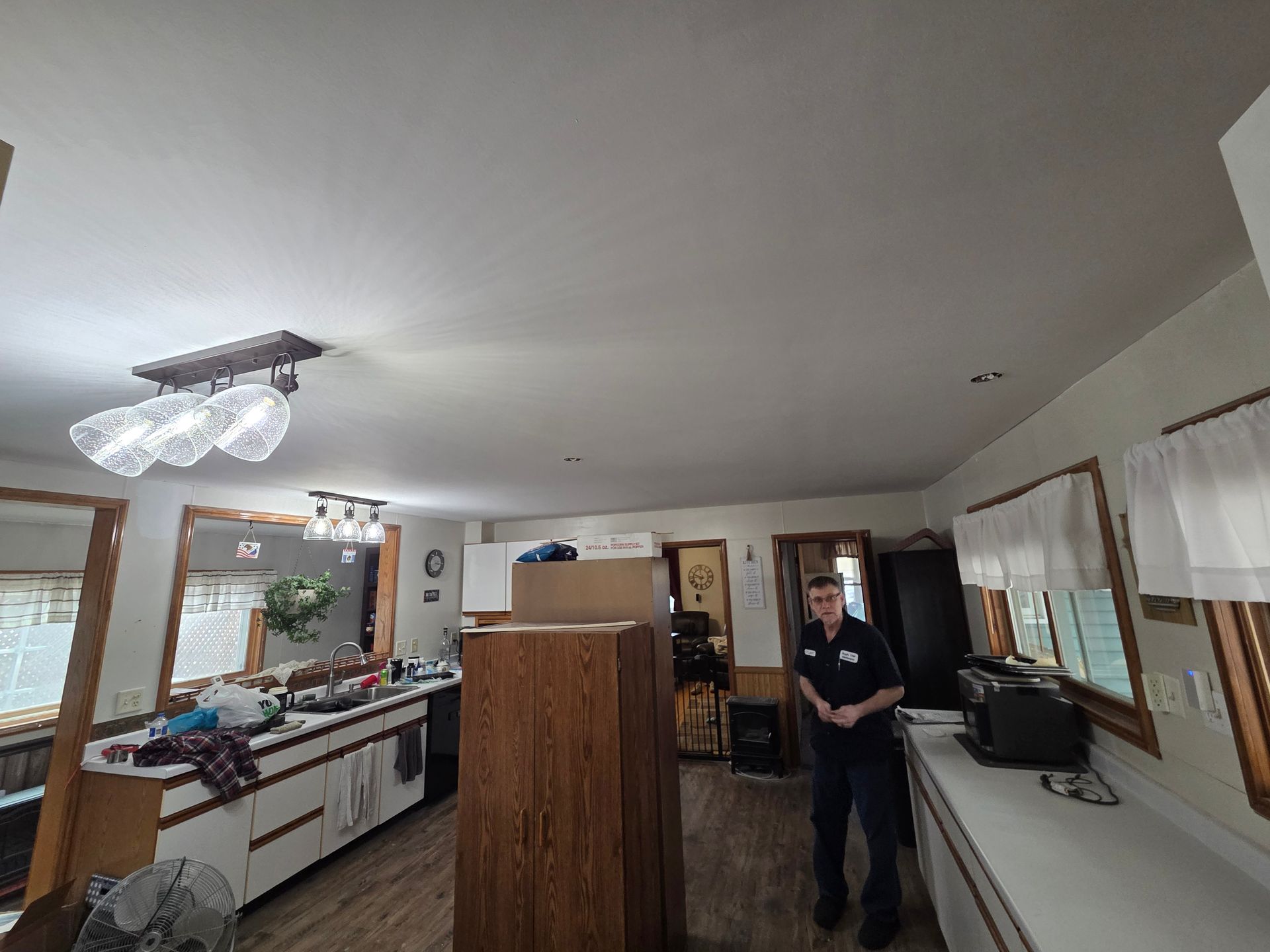 A person stands in a kitchen with a large wooden cabinet, white counters, and overhead light fixtures.