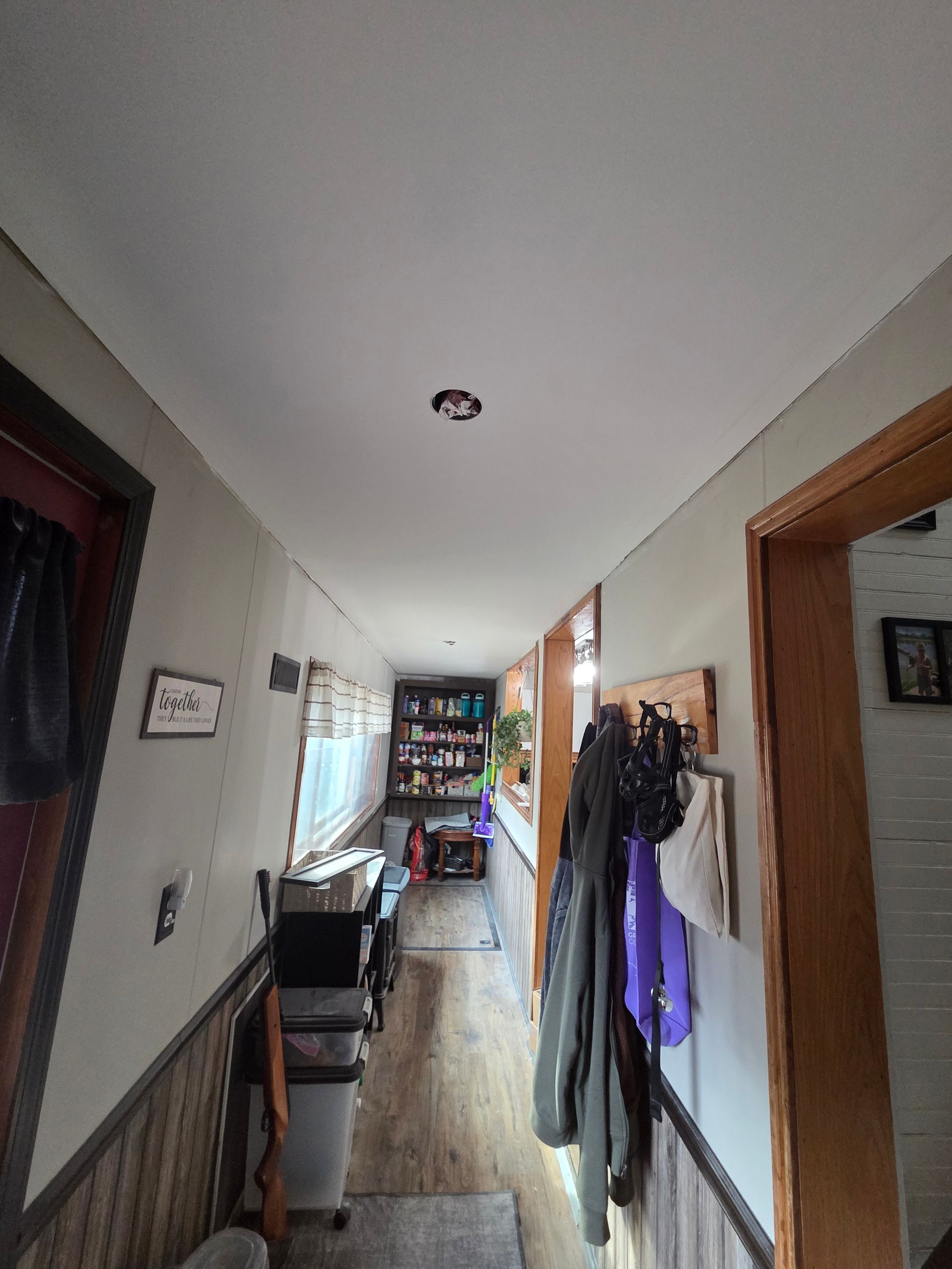 A narrow hallway with light gray walls, wood-paneled wainscoting, and a central ceiling light leading toward a cluttered area.