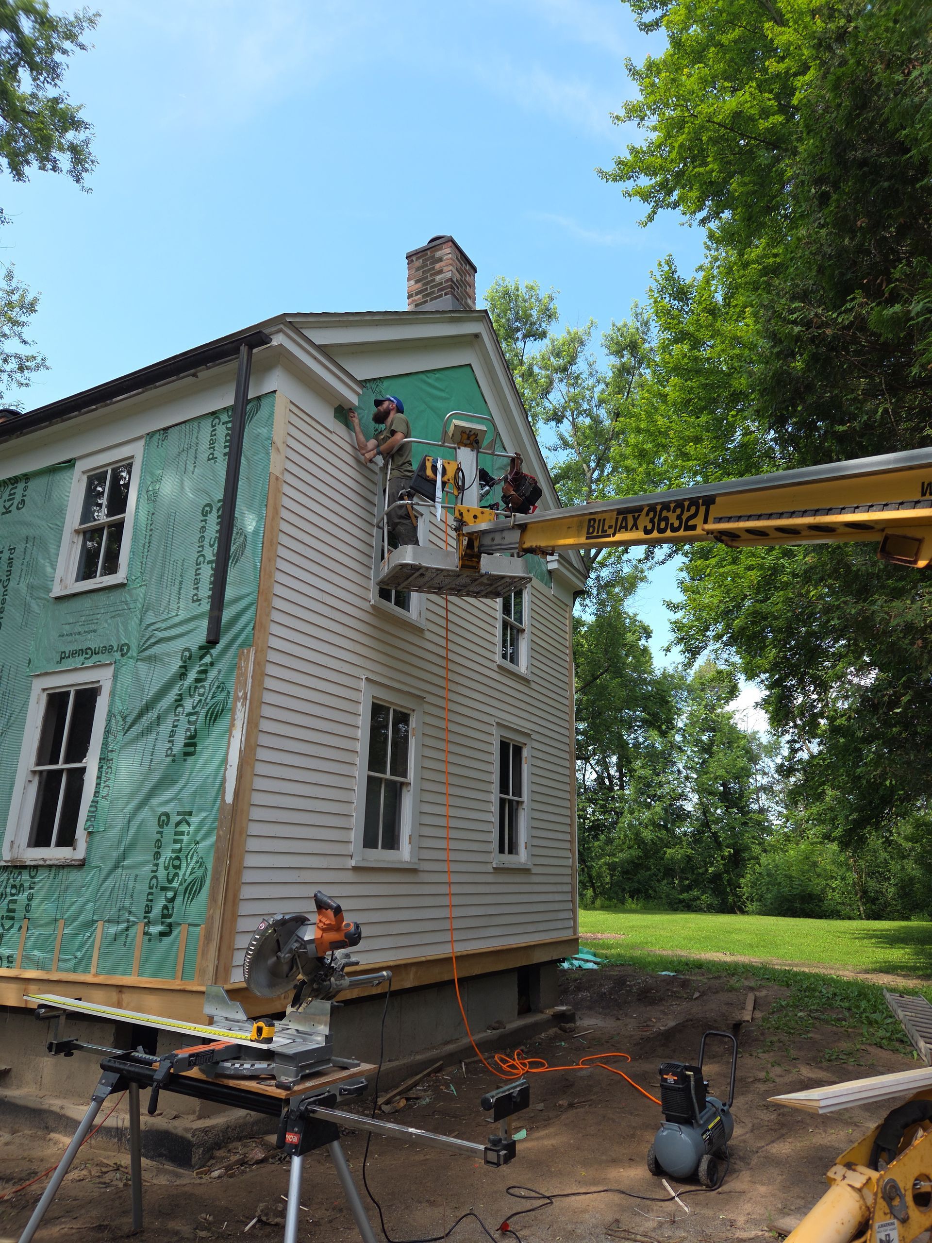 Workers on a boom lift replace siding on the exterior of a multi-story home under renovation on a sunny day.