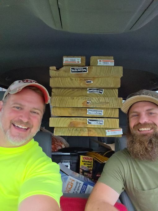 Two smiling people sit inside a vehicle with a stack of wooden boards secured between them and the roof.