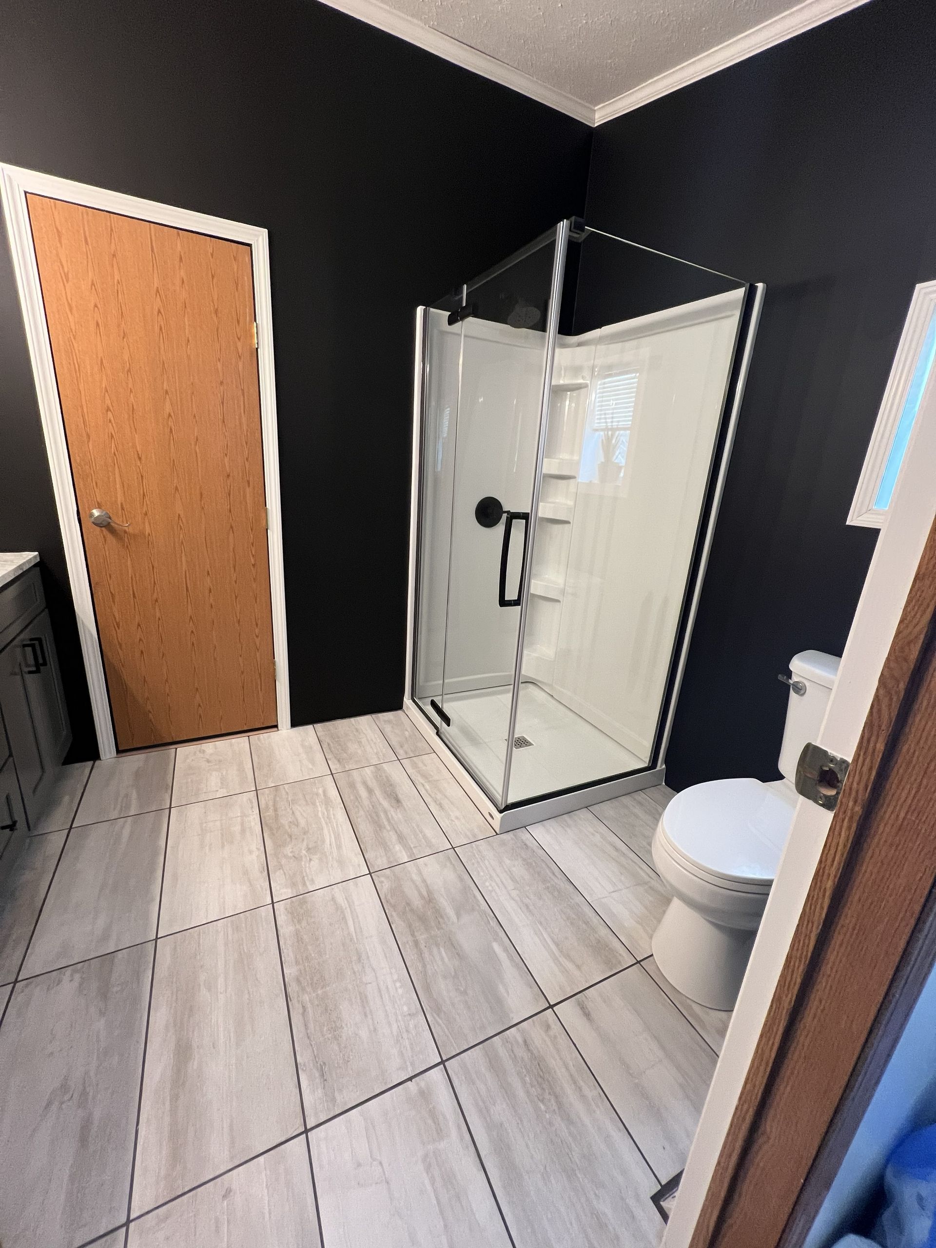 A bathroom with dark walls, light gray tile flooring, a white glass-enclosed shower, a toilet, and a wood-tone door.