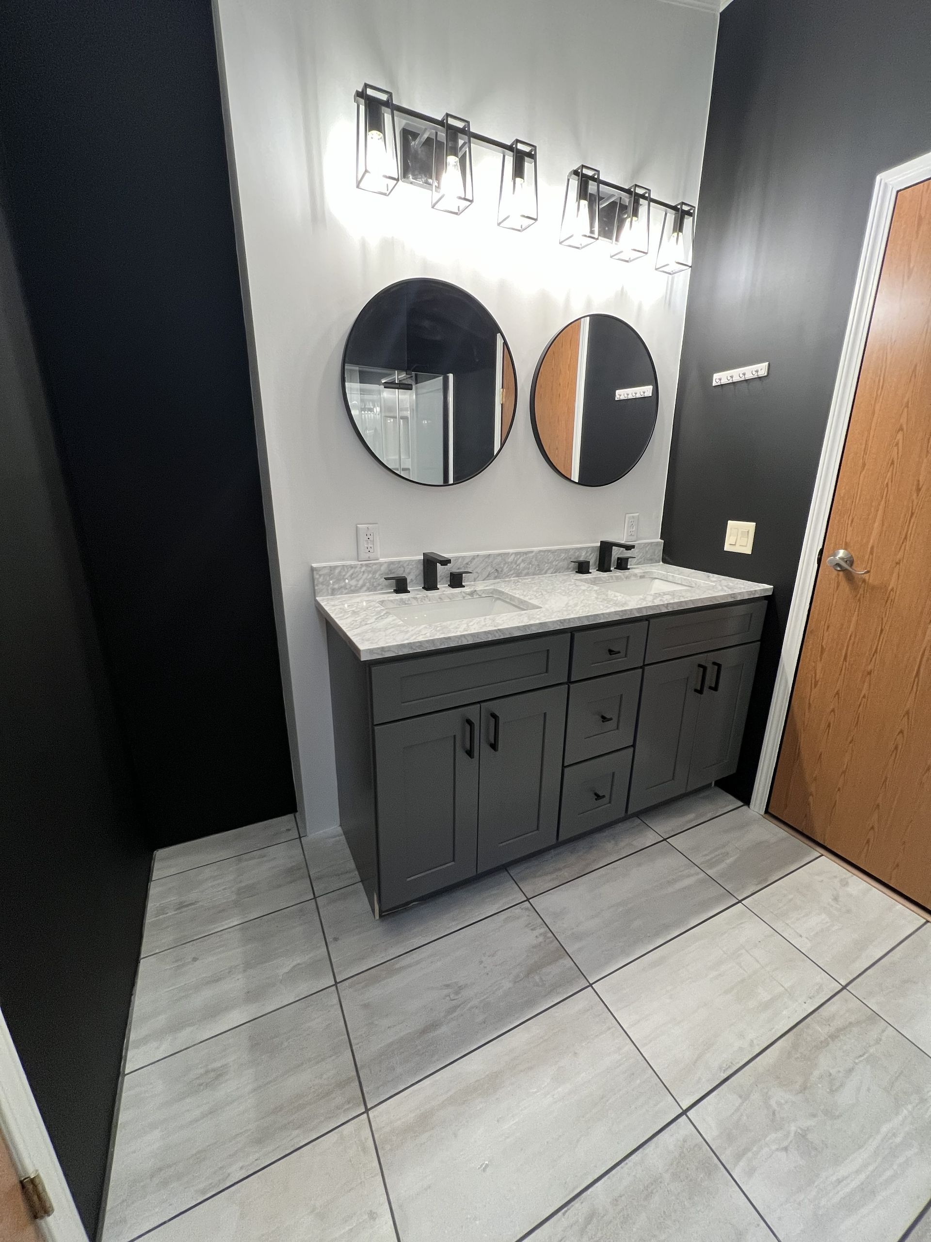 A bathroom with a double vanity featuring a grey cabinet, white countertop, two round mirrors, and black wall accents.