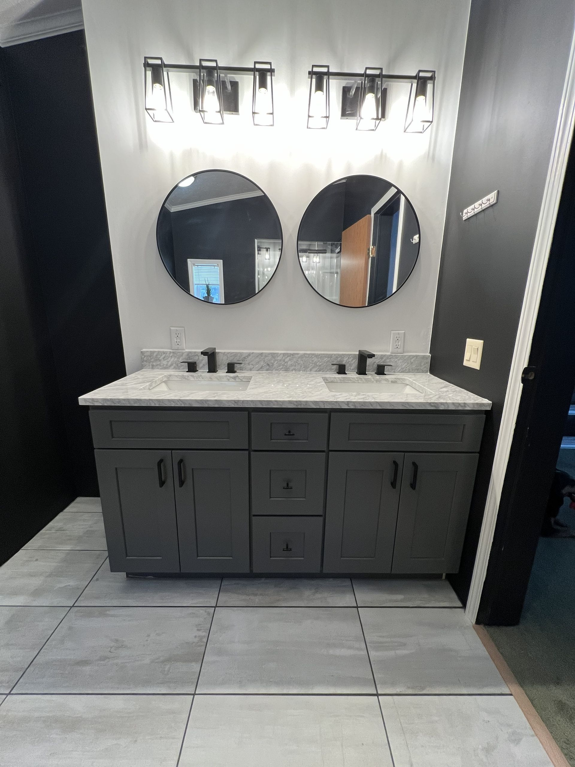 Double vanity bathroom sink with grey cabinets, light granite countertop, two round mirrors, and black light fixtures.