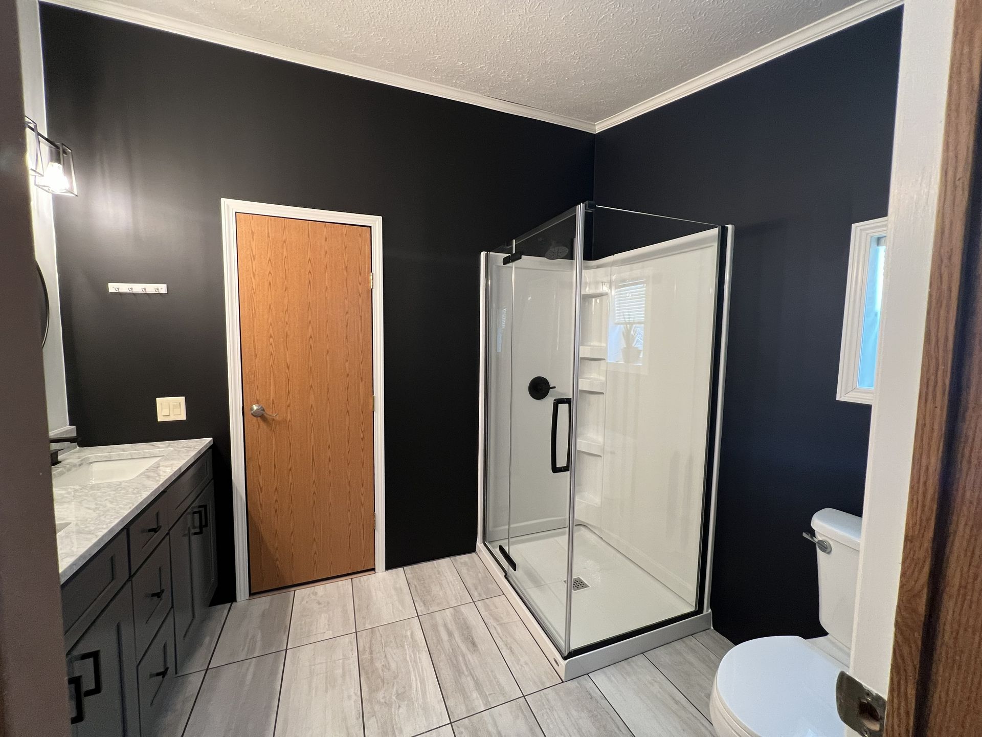 A modern bathroom featuring black walls, a wood-toned door, a glass shower, a marble-topped vanity, and a white toilet.