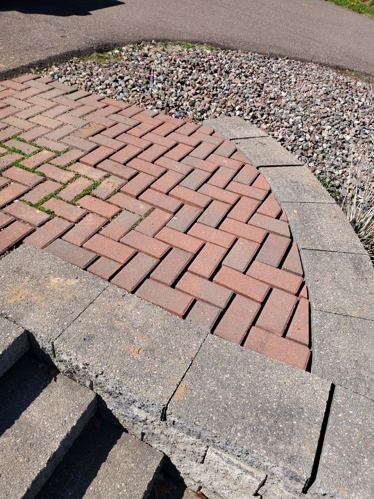 A curved brick patio bordered by grey concrete blocks, with stone steps in the foreground and a gravel area behind.