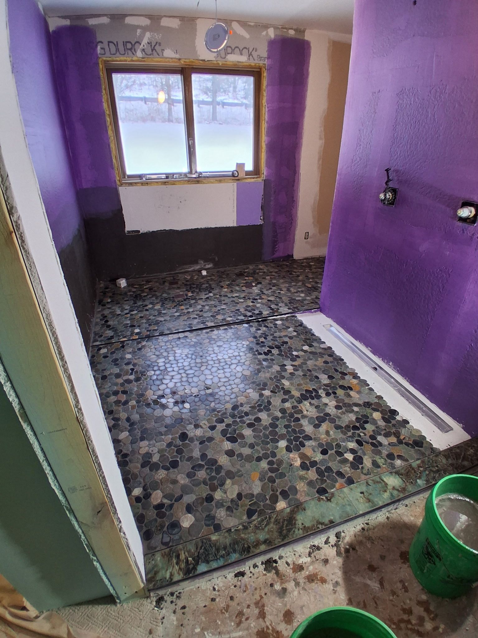 A bathroom renovation in progress, featuring a pebble-tiled shower floor and walls partially covered in purple waterproof membrane.