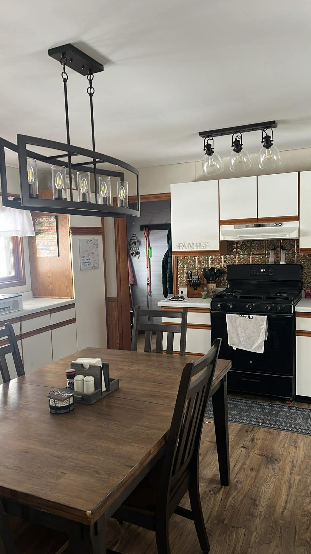 A dining table with a wooden top and black chairs sits in a kitchen featuring white cabinets and an industrial chandelier.