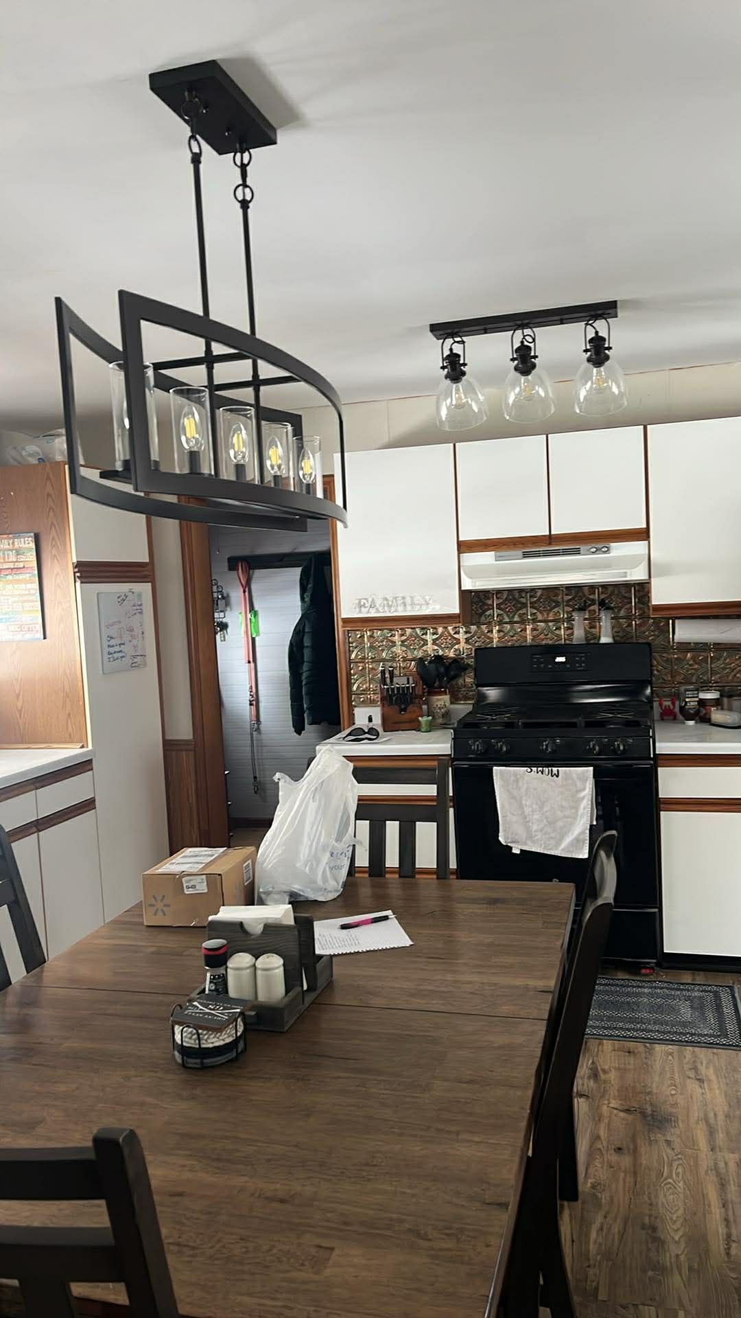 A wooden dining table sits beneath a modern industrial chandelier in a kitchen with white cabinets and a tile backsplash.