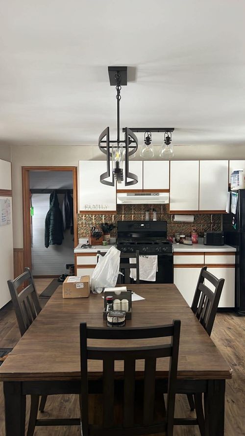 A wooden dining table with chairs sits in front of a white-cabinet kitchen featuring a dark pendant light and stove.