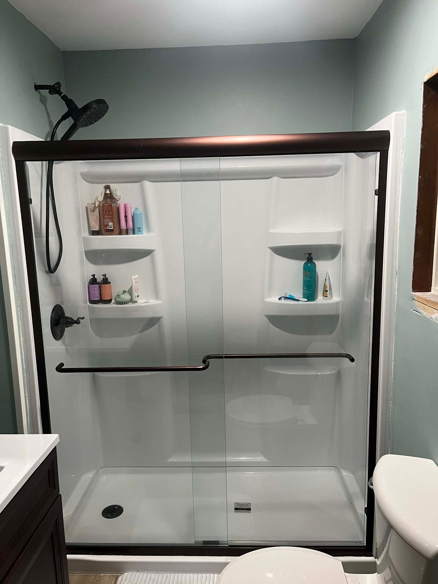 A walk-in shower with white walls, built-in shelving, a glass sliding door with a dark frame, and a handheld showerhead.