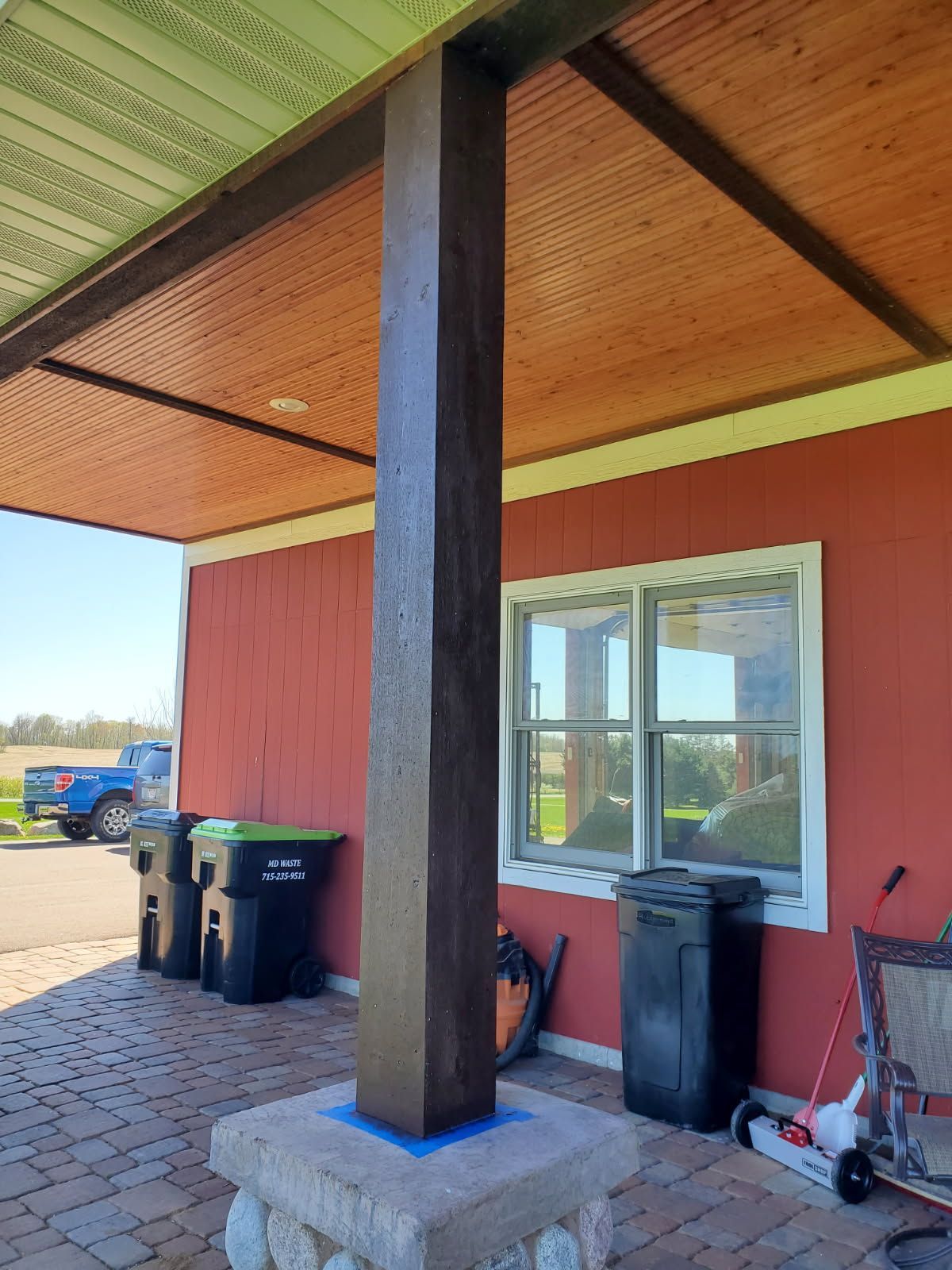 A dark brown wooden porch post sits on a square stone base in front of a red building with a wood-paneled ceiling.