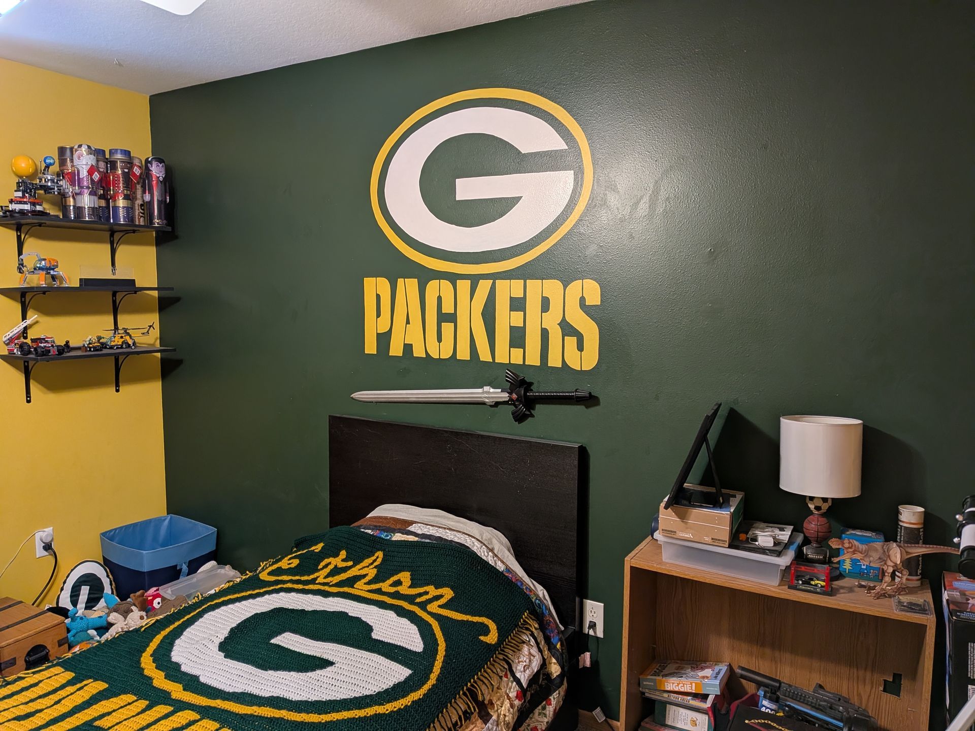 A Green Bay Packers-themed bedroom with a large logo on a dark green wall, a matching bedspread, and wall-mounted sword.