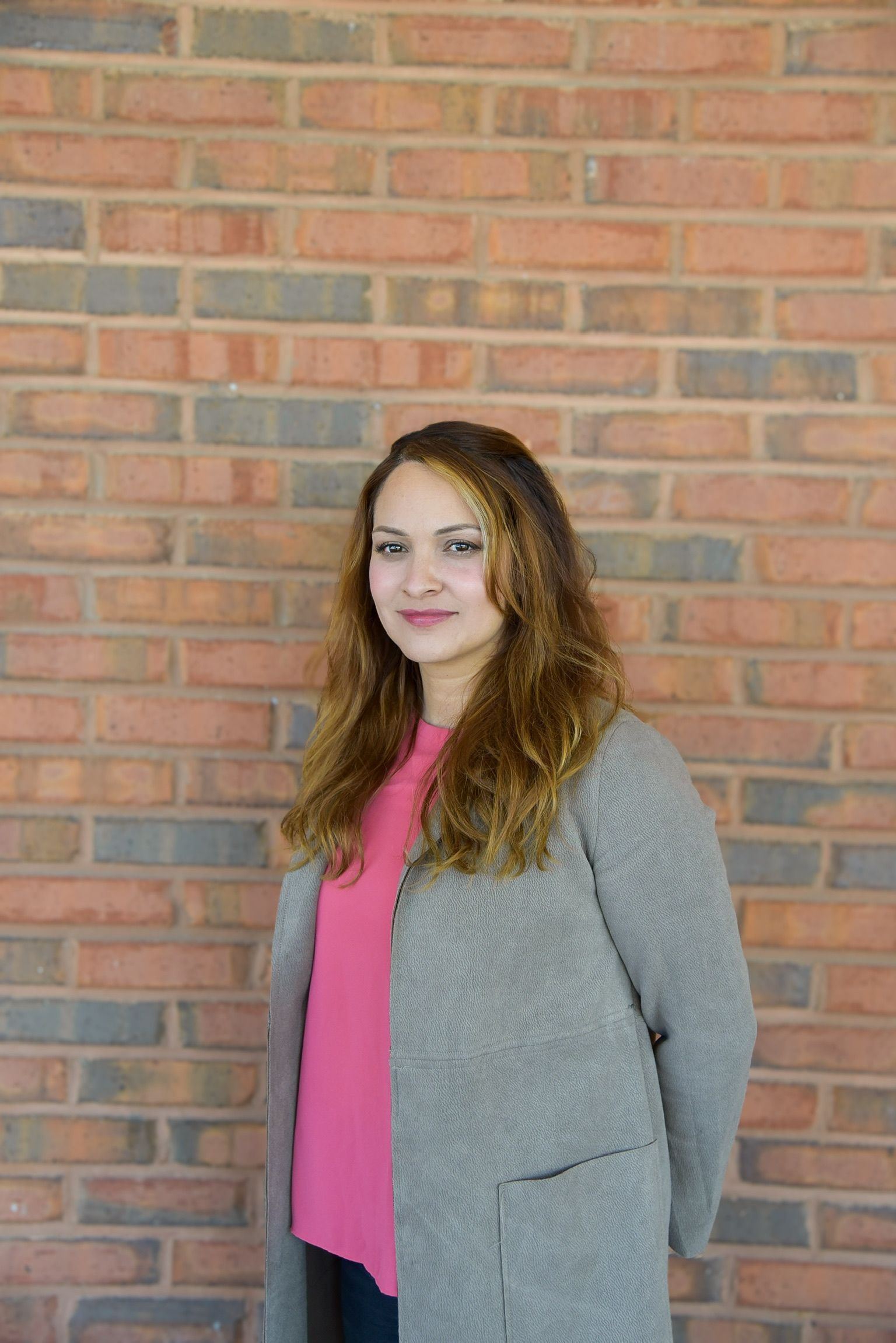 A woman in a pink shirt and gray coat is standing in front of a brick wall.