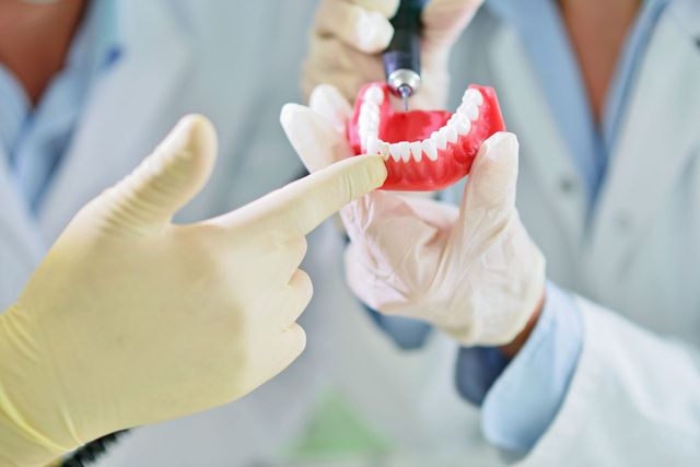 Un dentista mostra delle protesi dentarie a un paziente, con le mani guantate, in un ambiente clinico.