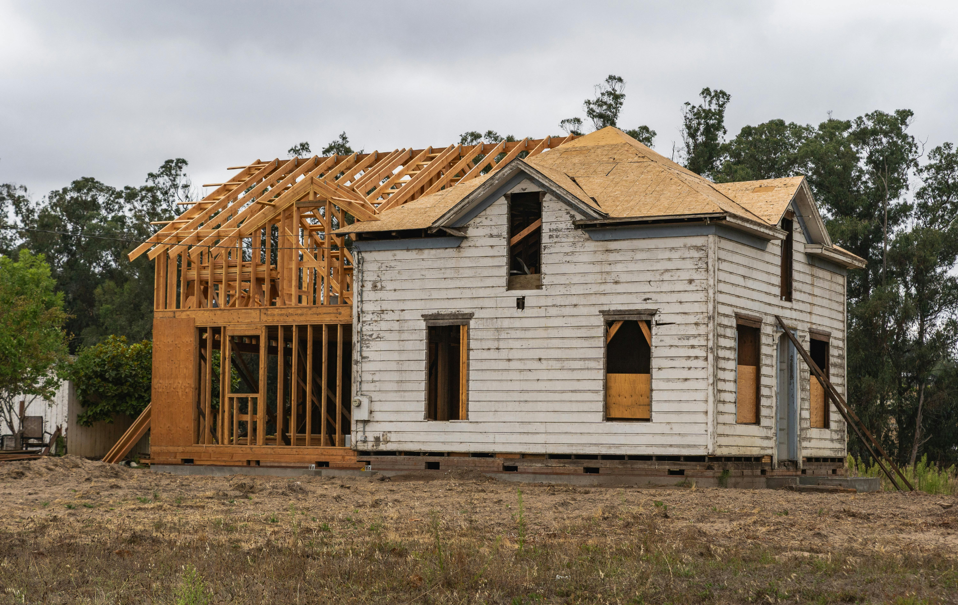 Old two-story house under renovation, with new wooden framework addition. Graying paint, overcast sky.