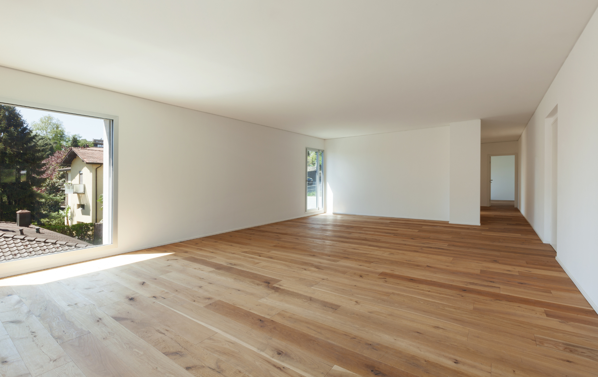 Empty room with wooden floors, white walls, and a large window overlooking trees.