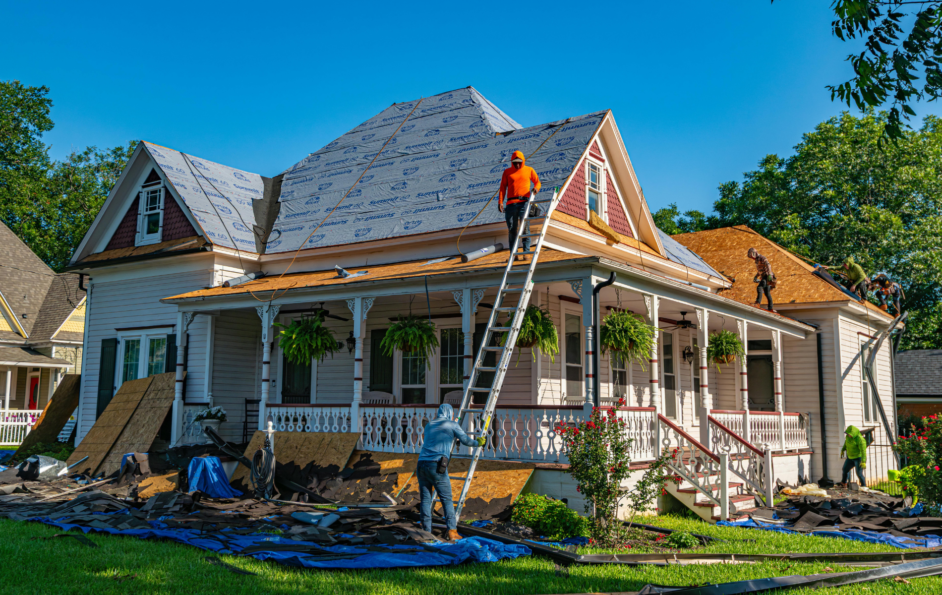 Workers replacing the roof of a two-story white house with a porch, blue tarp on the ground.