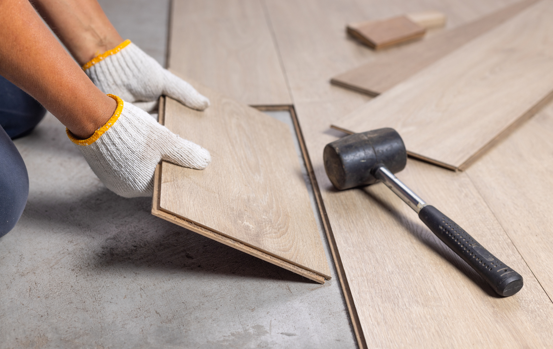 Person in gloves installing wooden floor planks on a concrete surface with a mallet nearby.