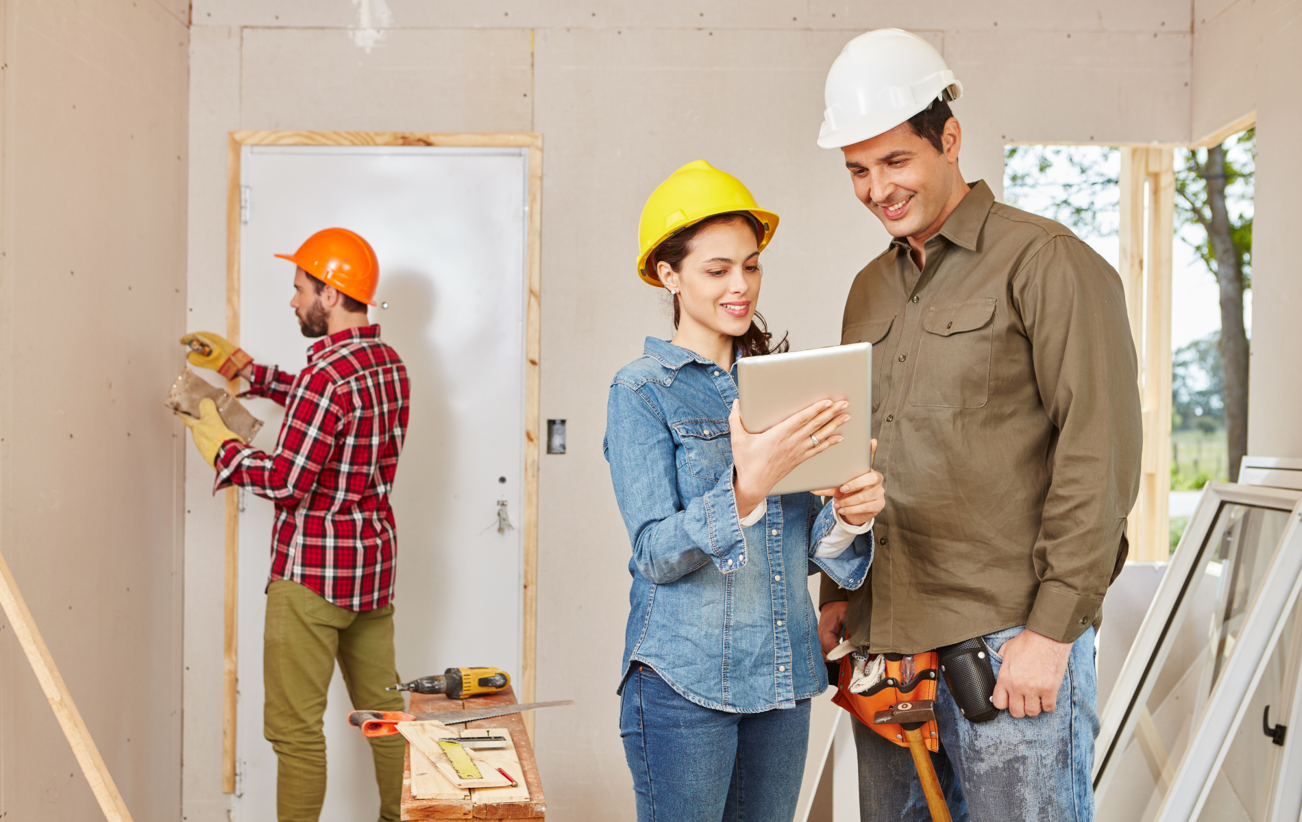 Three people in hard hats in a room under construction: one worker sanding a wall, two others reviewing a tablet.