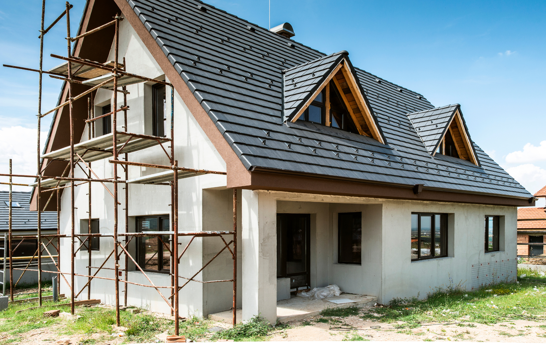 House under construction with scaffolding, gray stucco walls, dark roof tiles, and dormer windows.