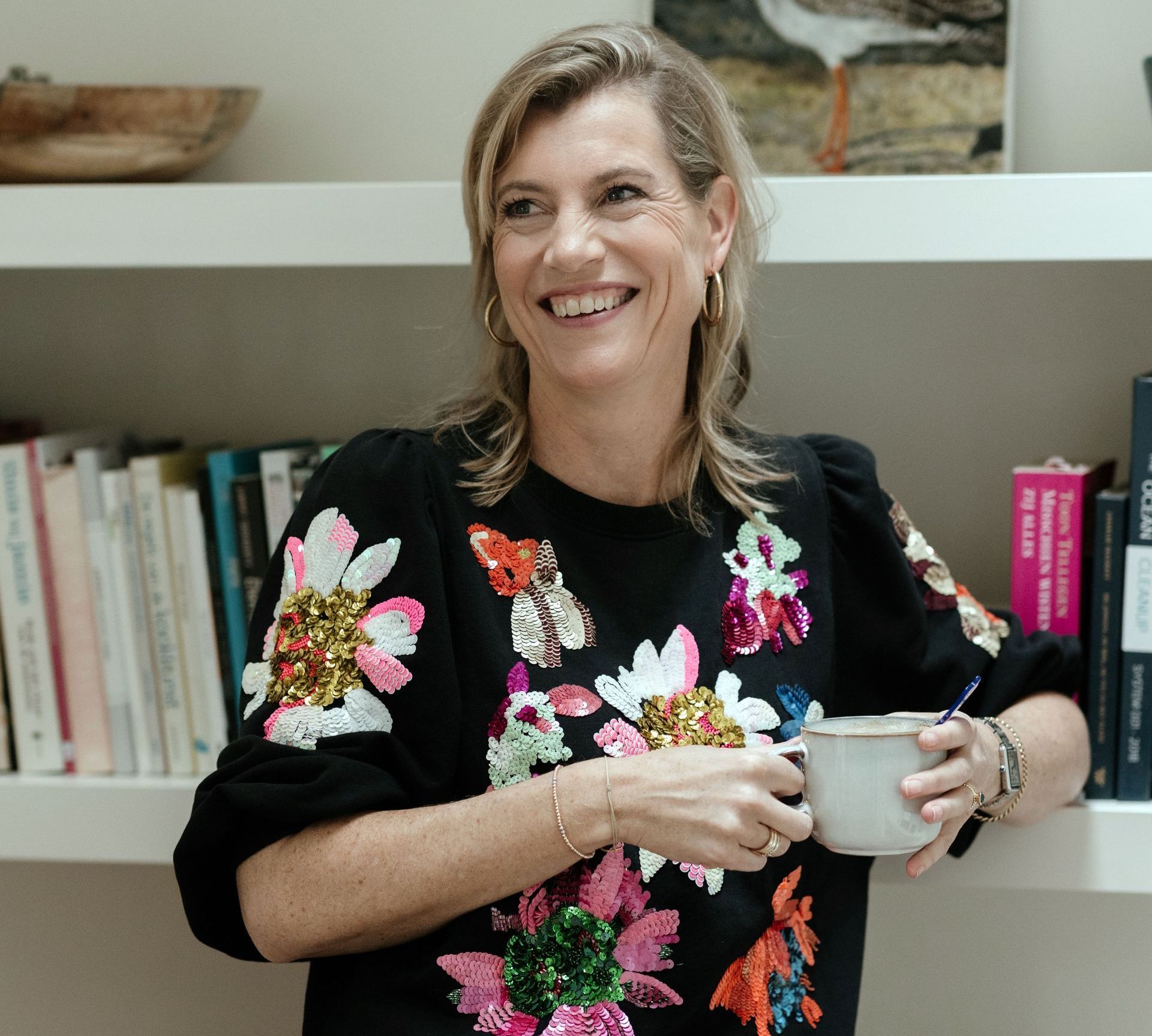 Een vrouw in een bloementrui houdt een kopje koffie vast en lacht.