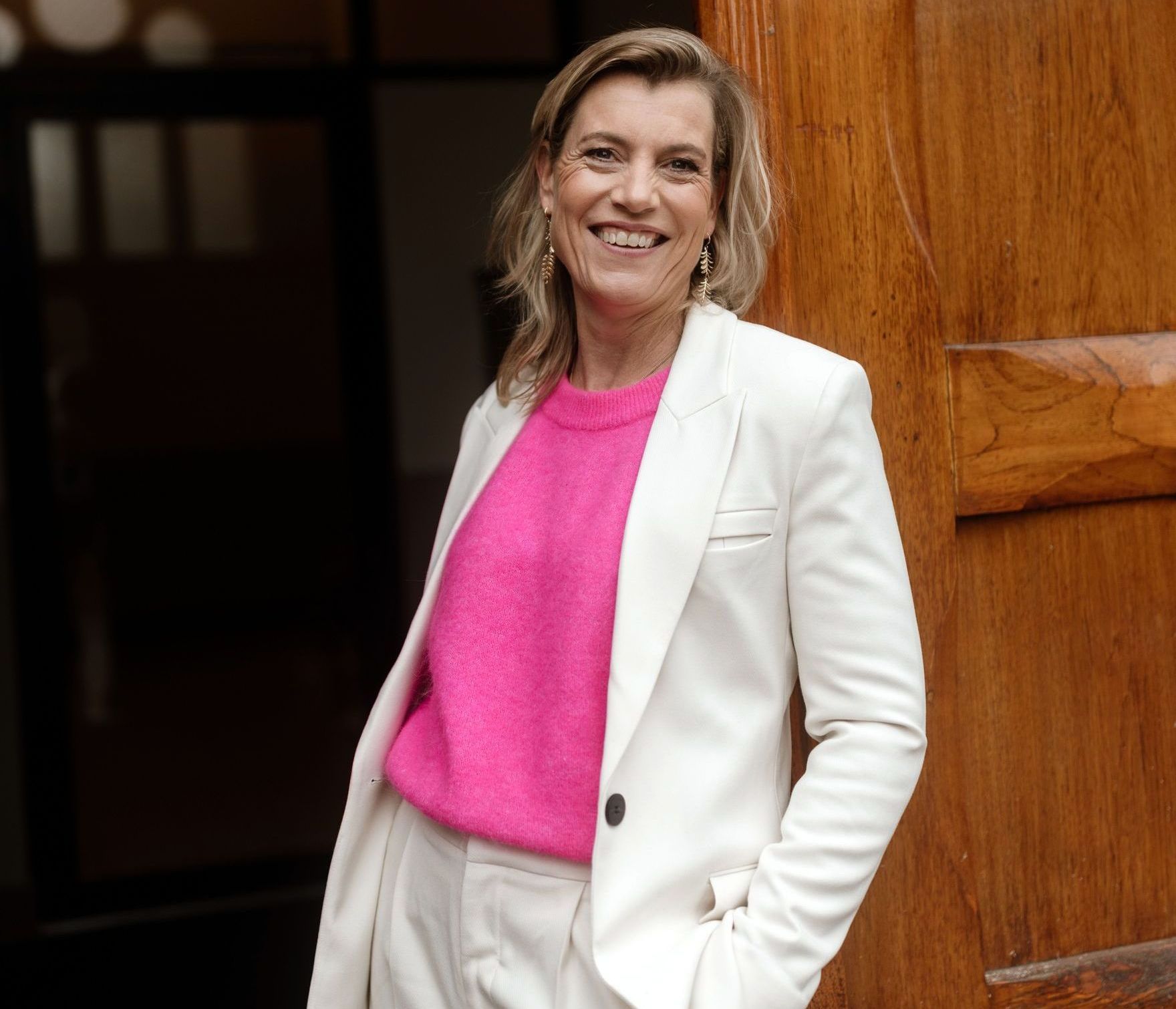 Een vrouw in een roze trui en een wit pak praat op een mobiele telefoon.