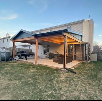 Covered backyard patio with a wooden roof, seating area, and string lights beside a house at dusk
