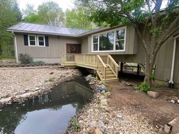 Single-story house with a small wooden porch over a rocky creek or pond in a wooded yard