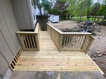 Wooden accessibility ramp and deck beside a house, with railings and a dirt yard beyond.