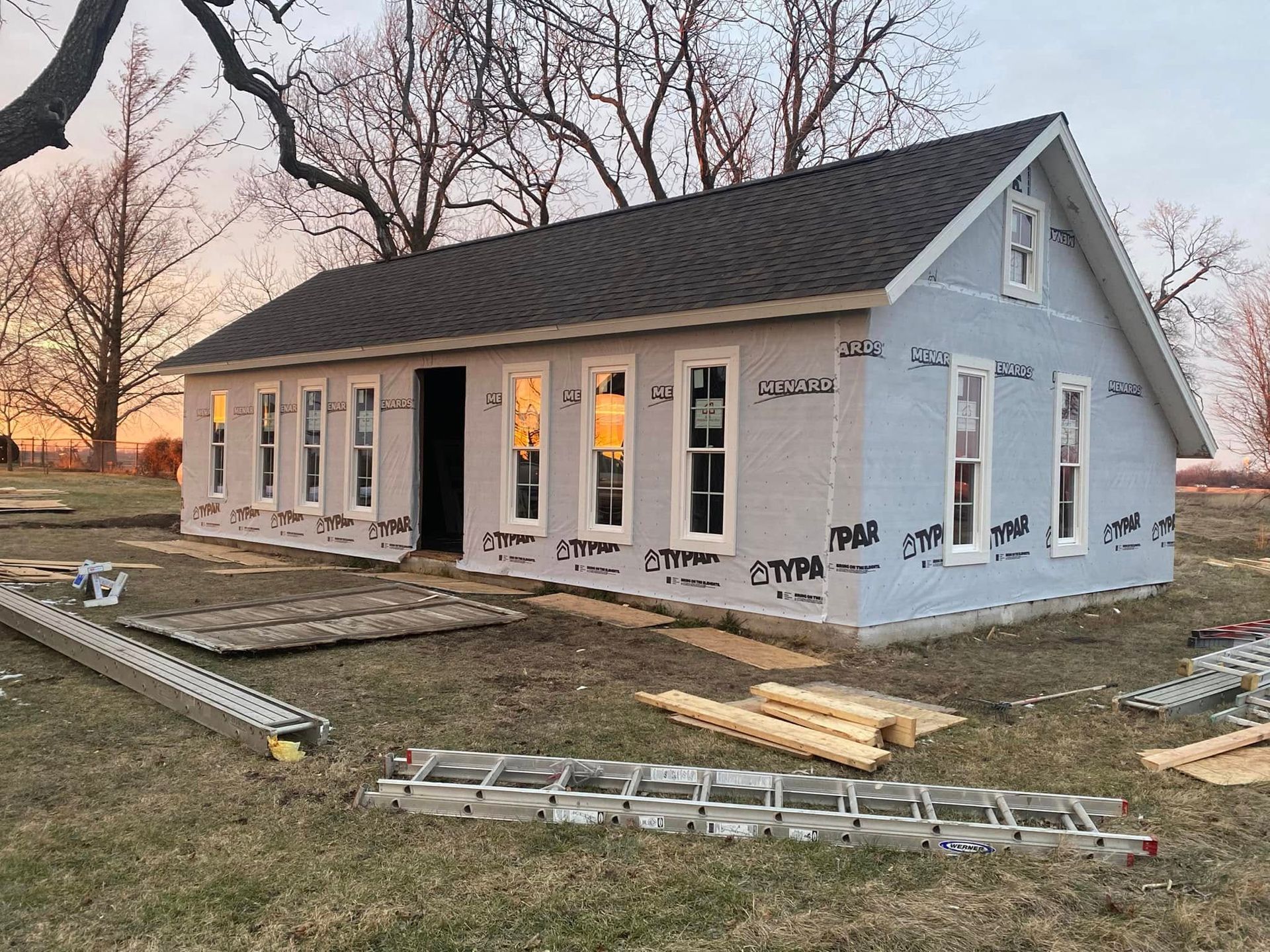 Partially built small house with white siding, black roof, and windows at sunset on a construction site