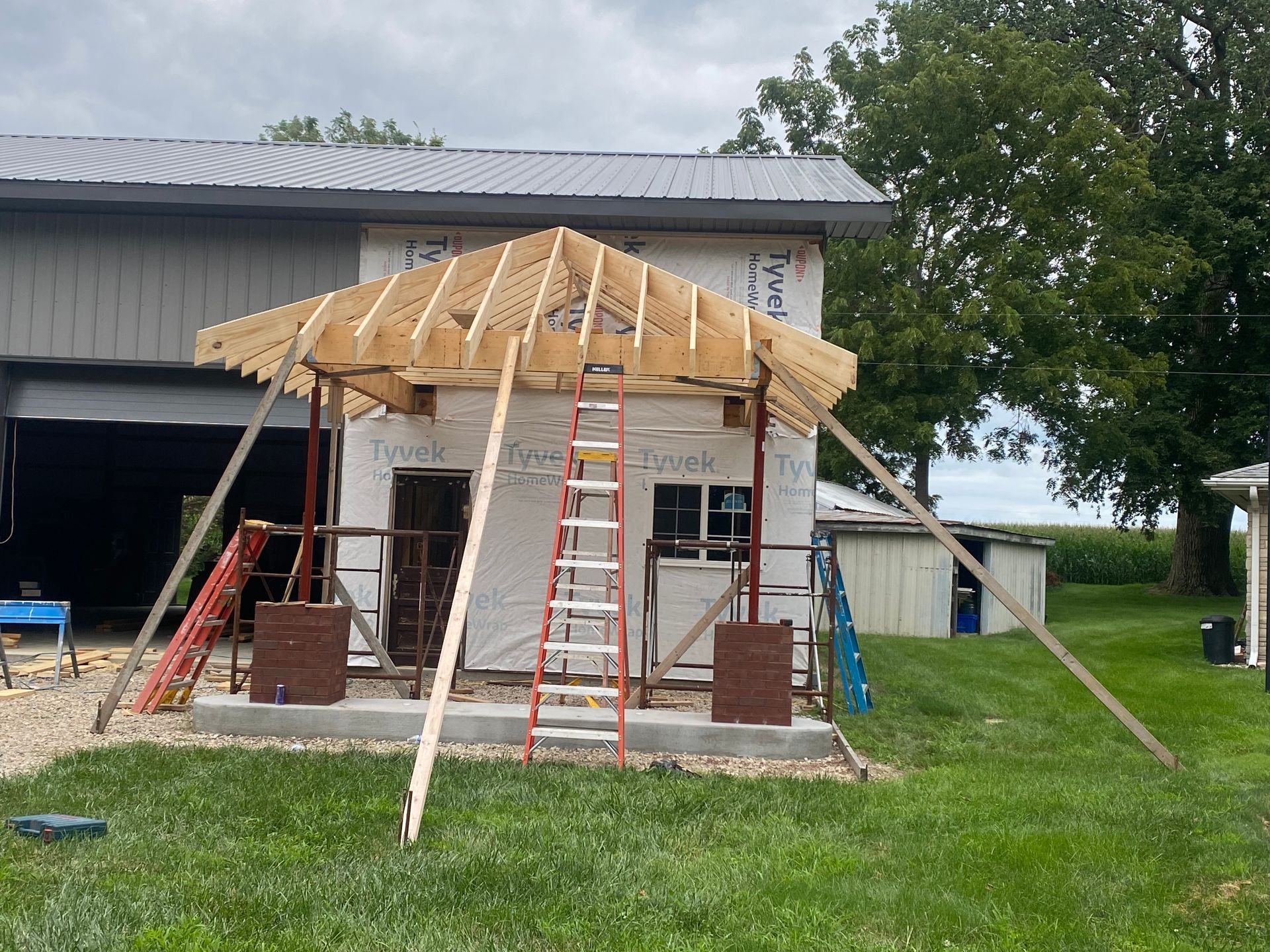 Front porch under construction with exposed framing, ladder, tools, and siding on a sunny day.