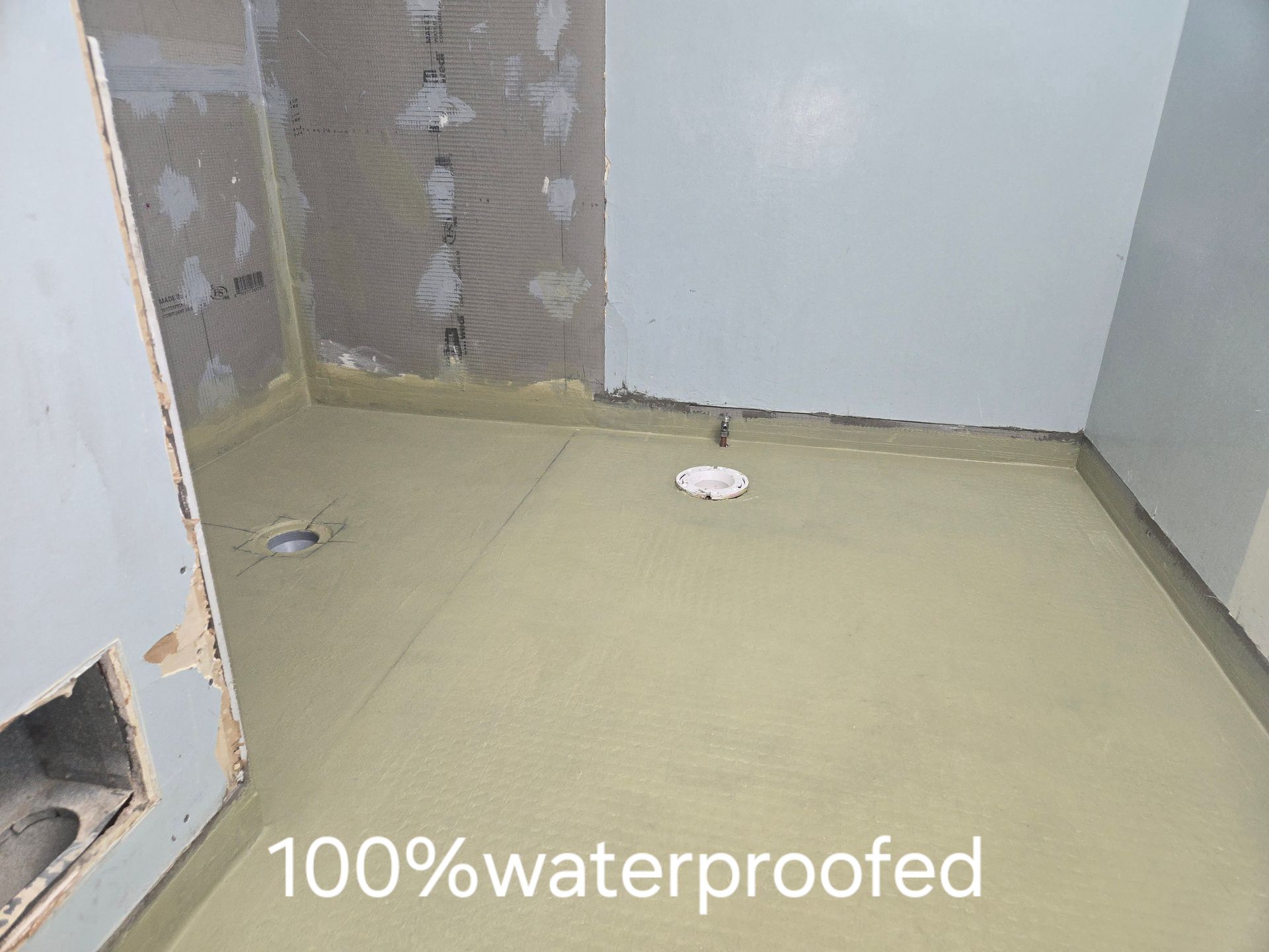 A person applying blue waterproof sealant to a corner of a concrete room with a brush, near a bucket.