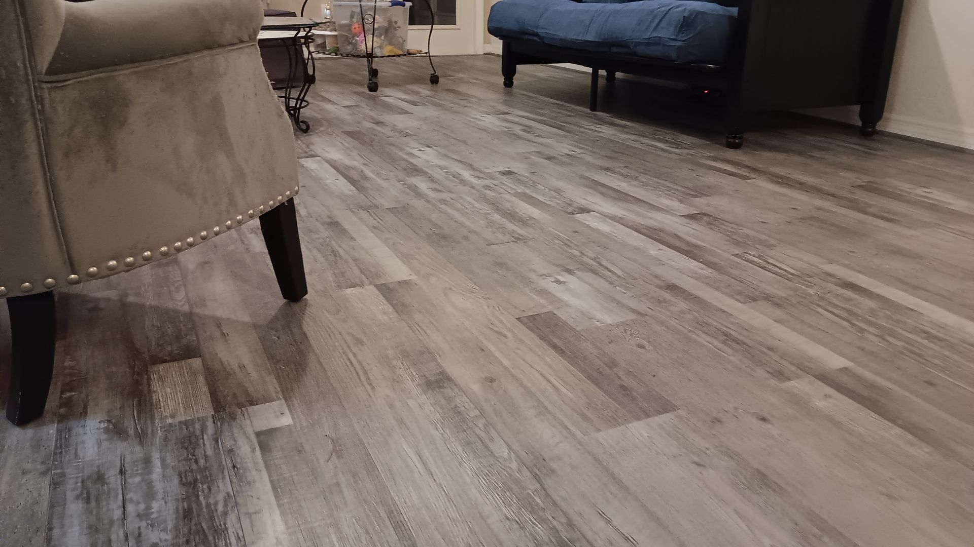 Close-up of gray wood-look floor with a tan armchair on the left and a bed and table in the background.