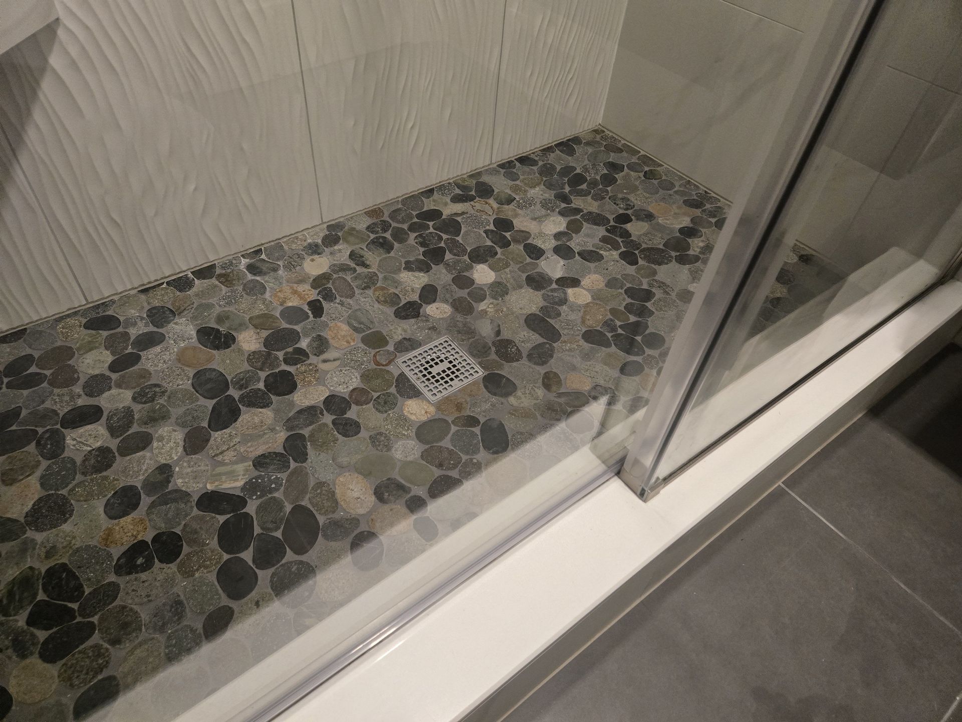 Shower with pebble floor, glass door, and white tiled walls.