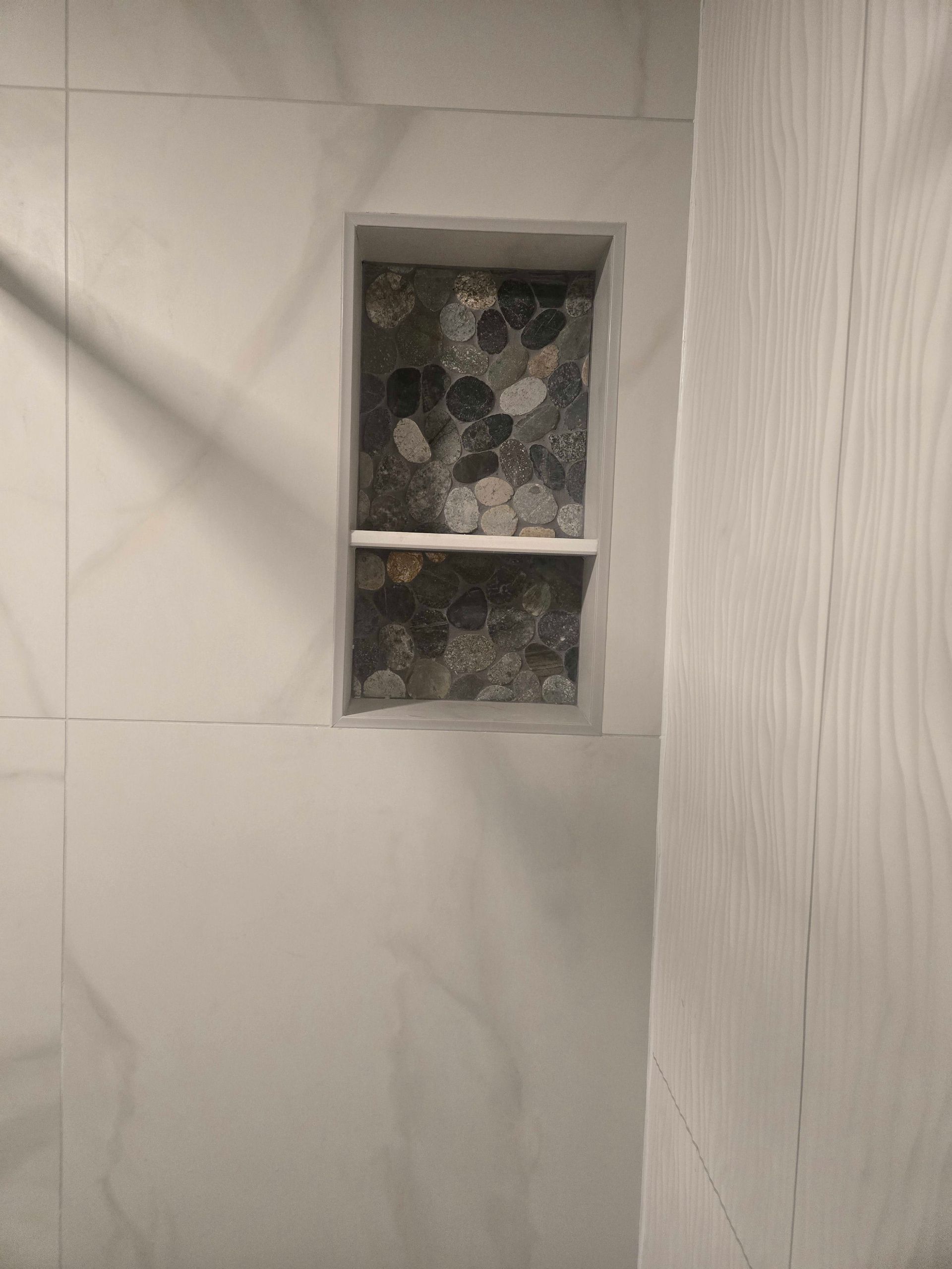 Built-in shower niche with pebble tile backing, white tile surround, and a shelf.
