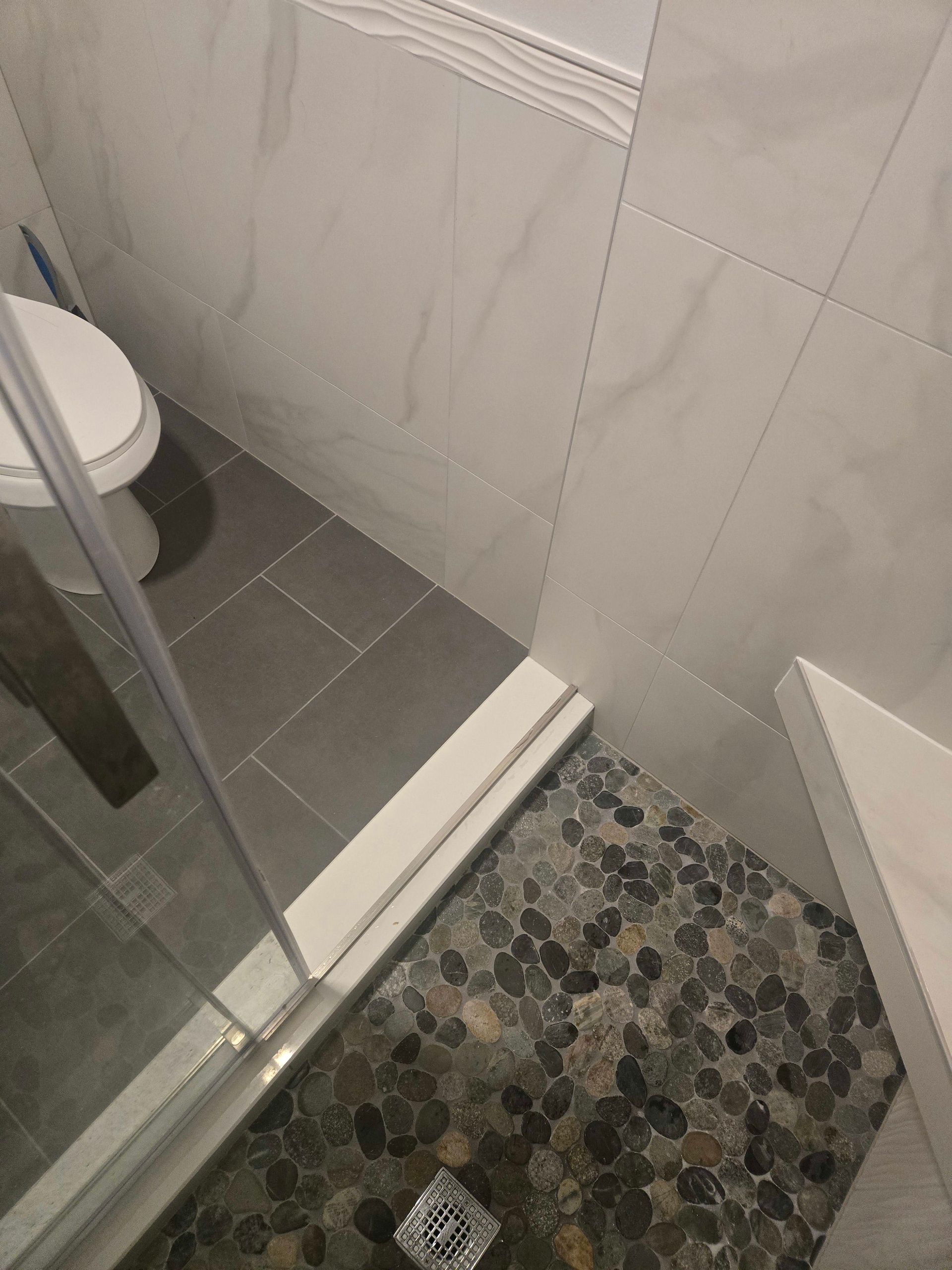 Shower with pebble floor, gray tile walls, and a glass door.