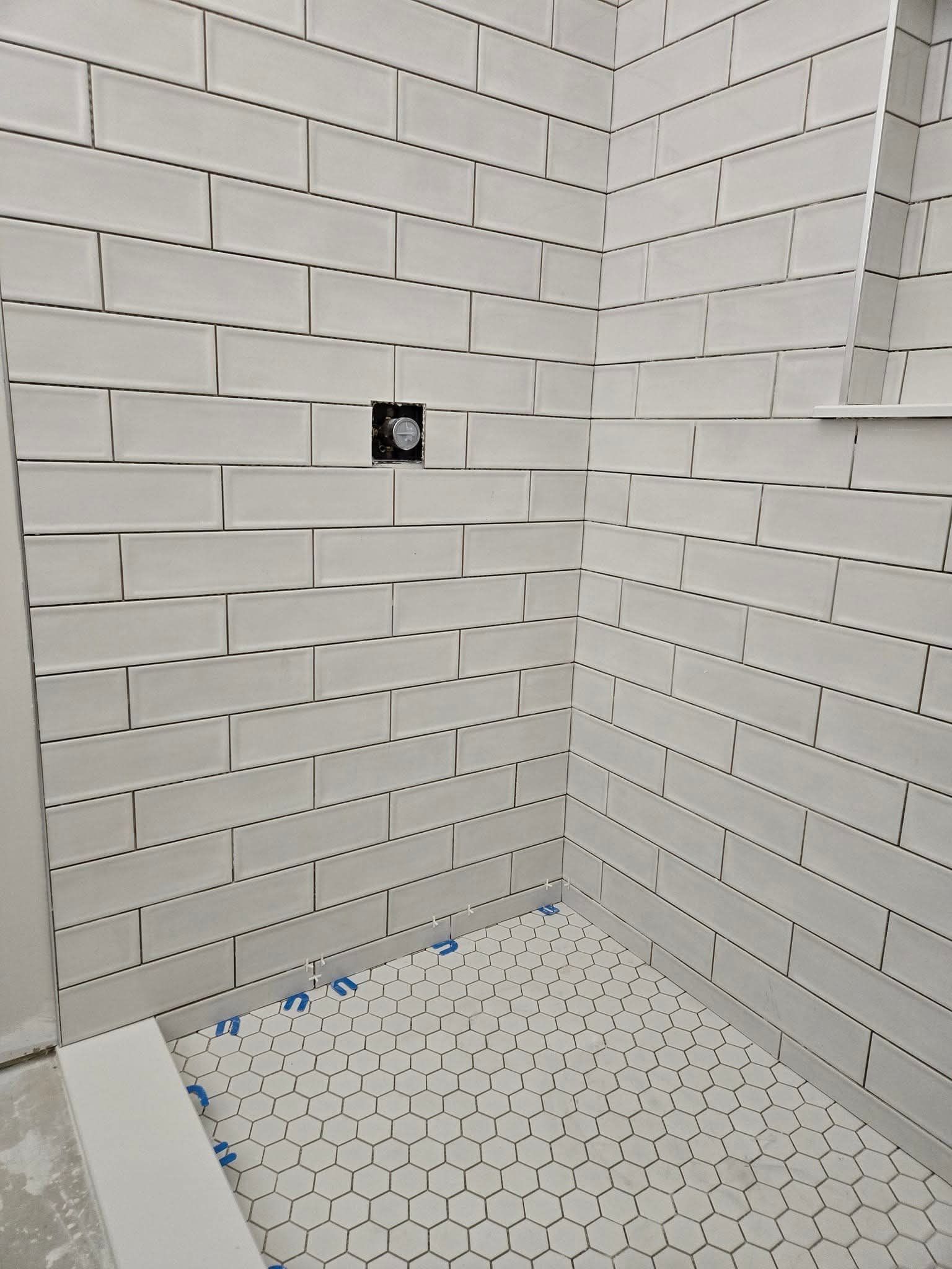 White tiled shower with black grout, featuring hexagon floor tile and a plumbing fixture.