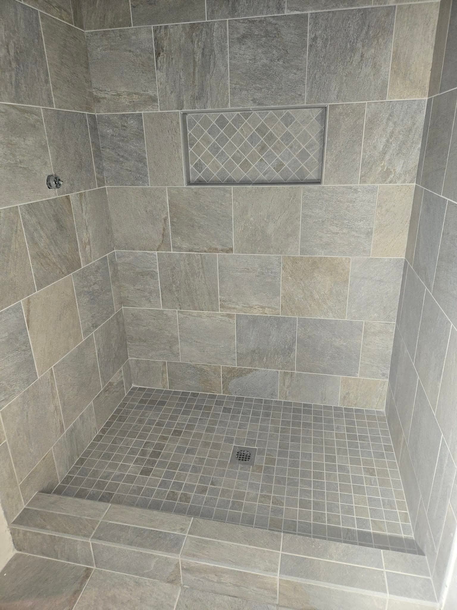 Tiled shower stall with a built-in recessed shelf. Grey stone tiles cover the walls and floor.