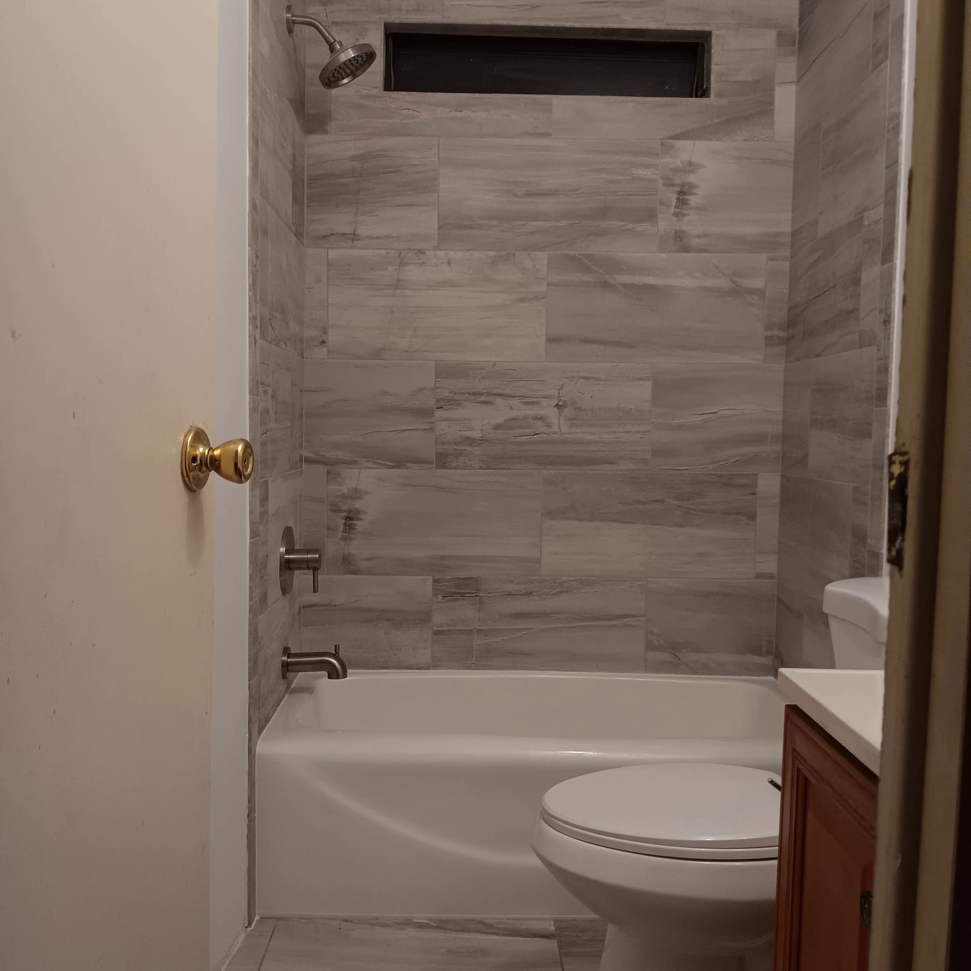 Bathroom with gray tiled shower wall, white tub, and toilet. A window is above the showerhead.