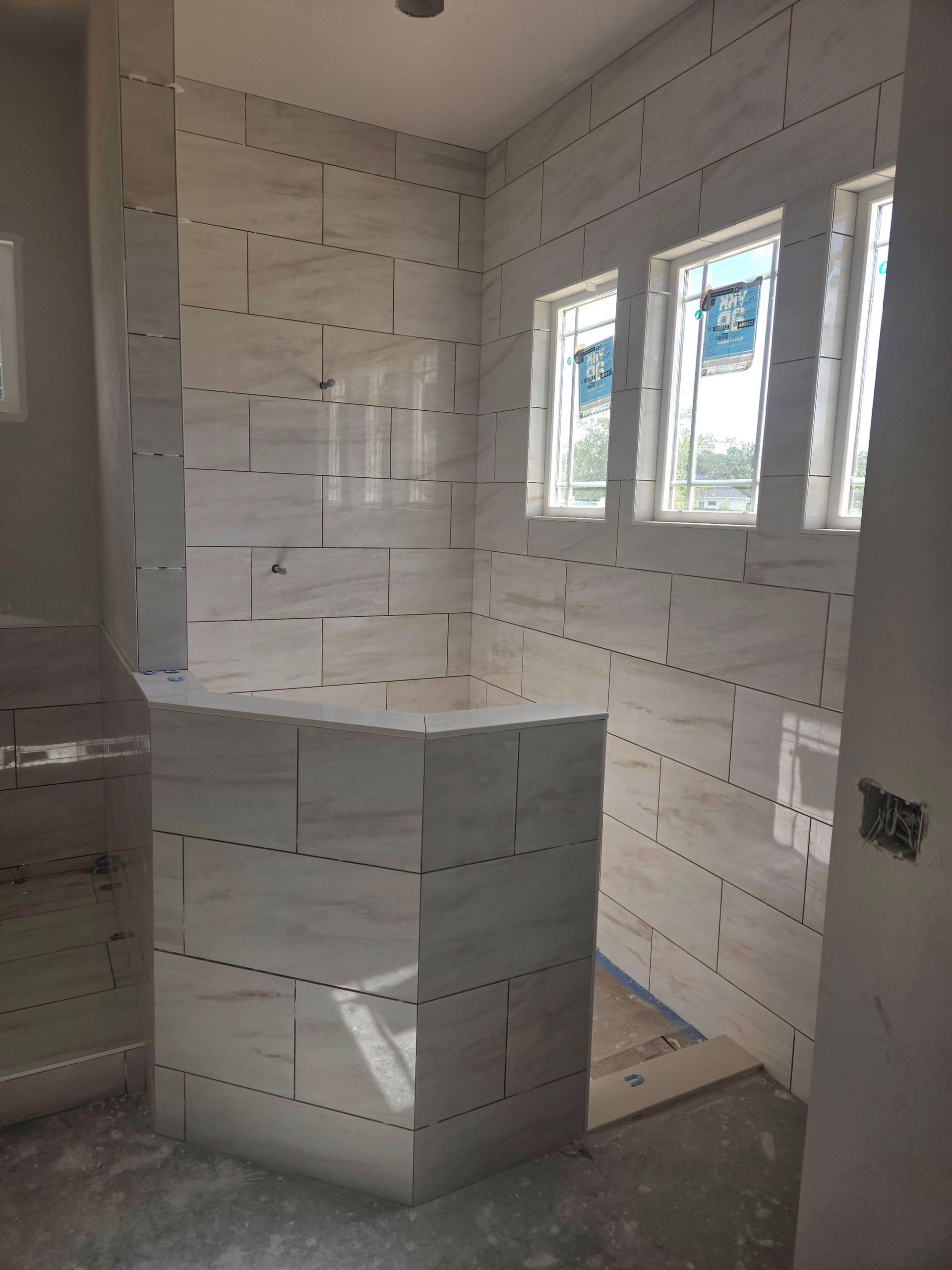 Bathroom with white marble tile on walls and a half-wall, three windows with blue wrapping, and unfinished flooring.