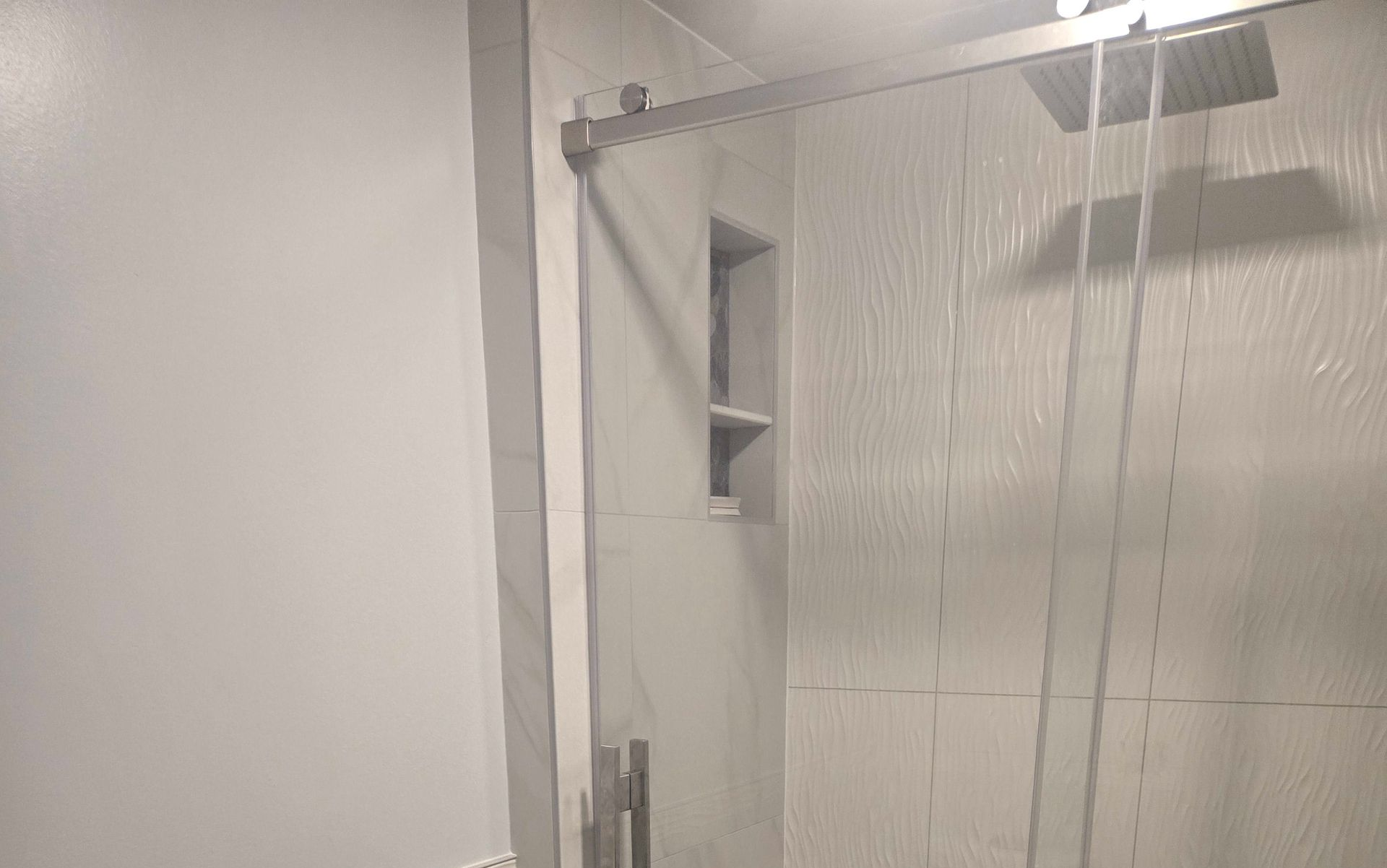 Shower with clear glass door, white textured tile, recessed shelf, and silver fixtures.