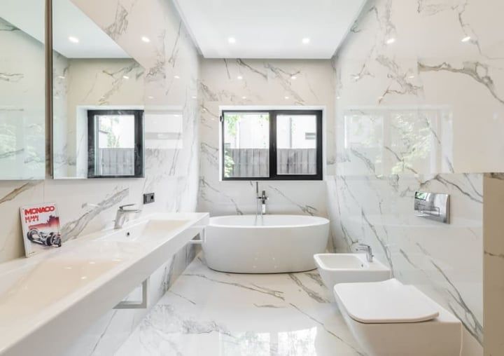 Modern bathroom with marble walls and floors, featuring a bathtub, sink, toilet, and windows.