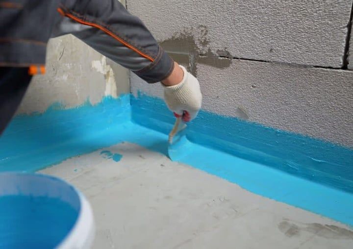 Person applying blue waterproof coating to a corner wall and floor with a brush.