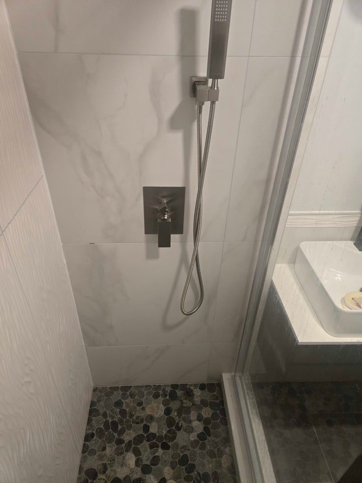 Shower with pebble floor, marble-look tiled walls, and brushed metal shower fixtures.  A clear glass shower door is visible.