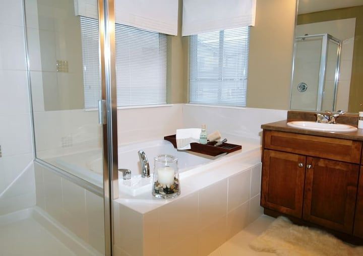 Bathroom with a white tiled tub, glass shower, wooden vanity, and a window with blinds.